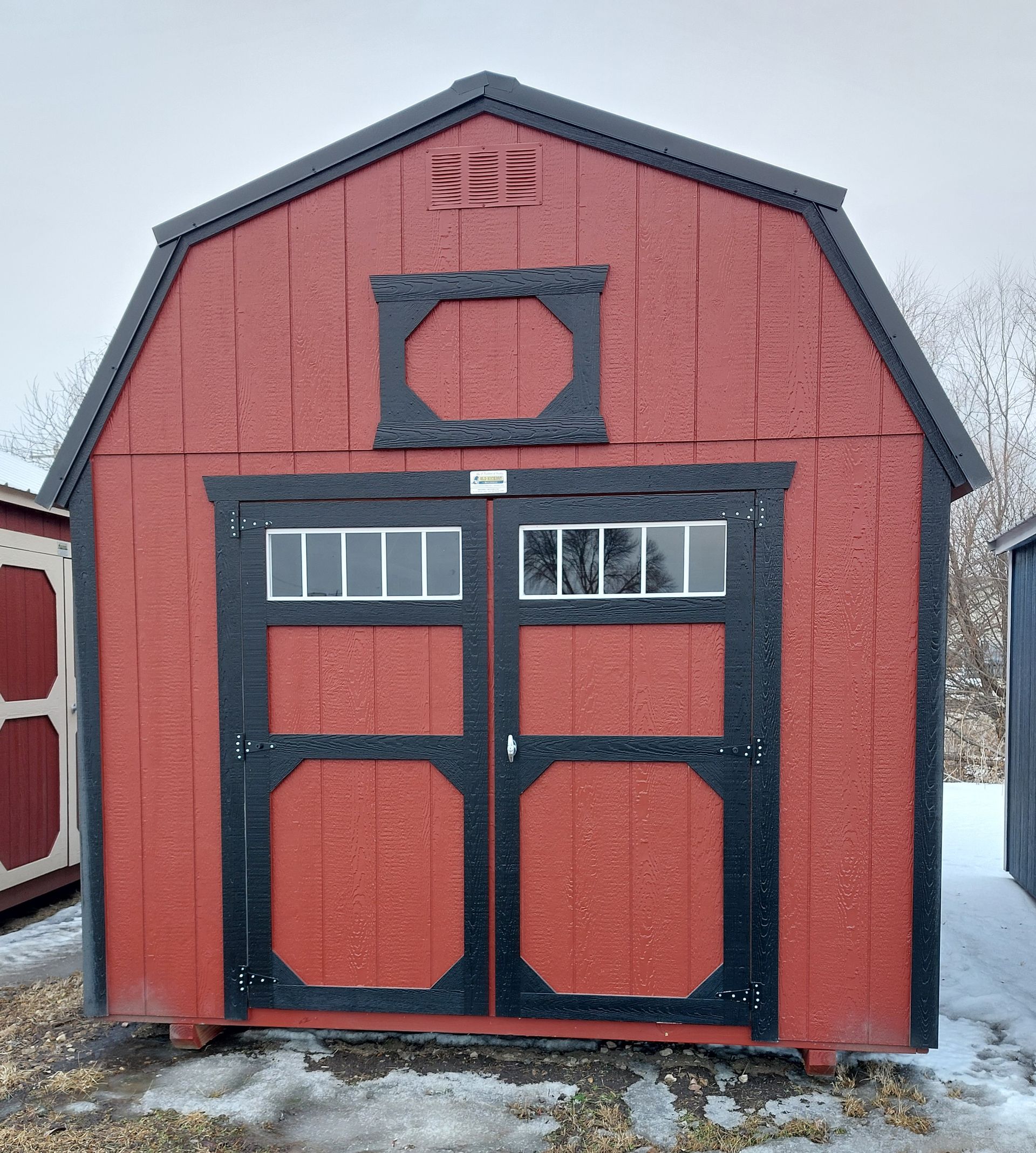 A red wooden shed with a gambrel roof and black trim, featuring double doors with windows, standing in a snowy field.