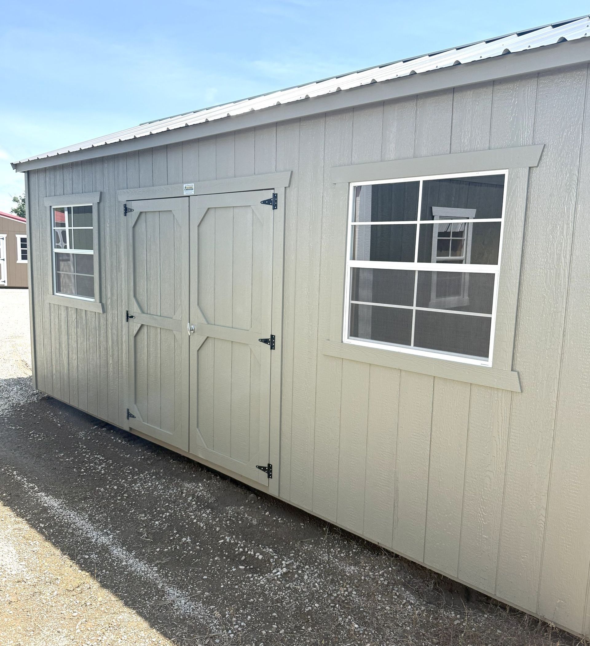 A beige storage shed with a metal roof, double doors, and two windows, parked on a gravel lot under a blue sky.
