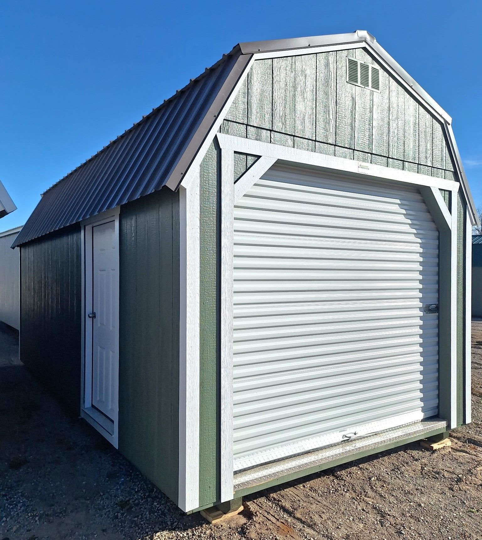 A dark green storage shed with a white roll-up garage door, a side entry door, and a metal gambrel roof.