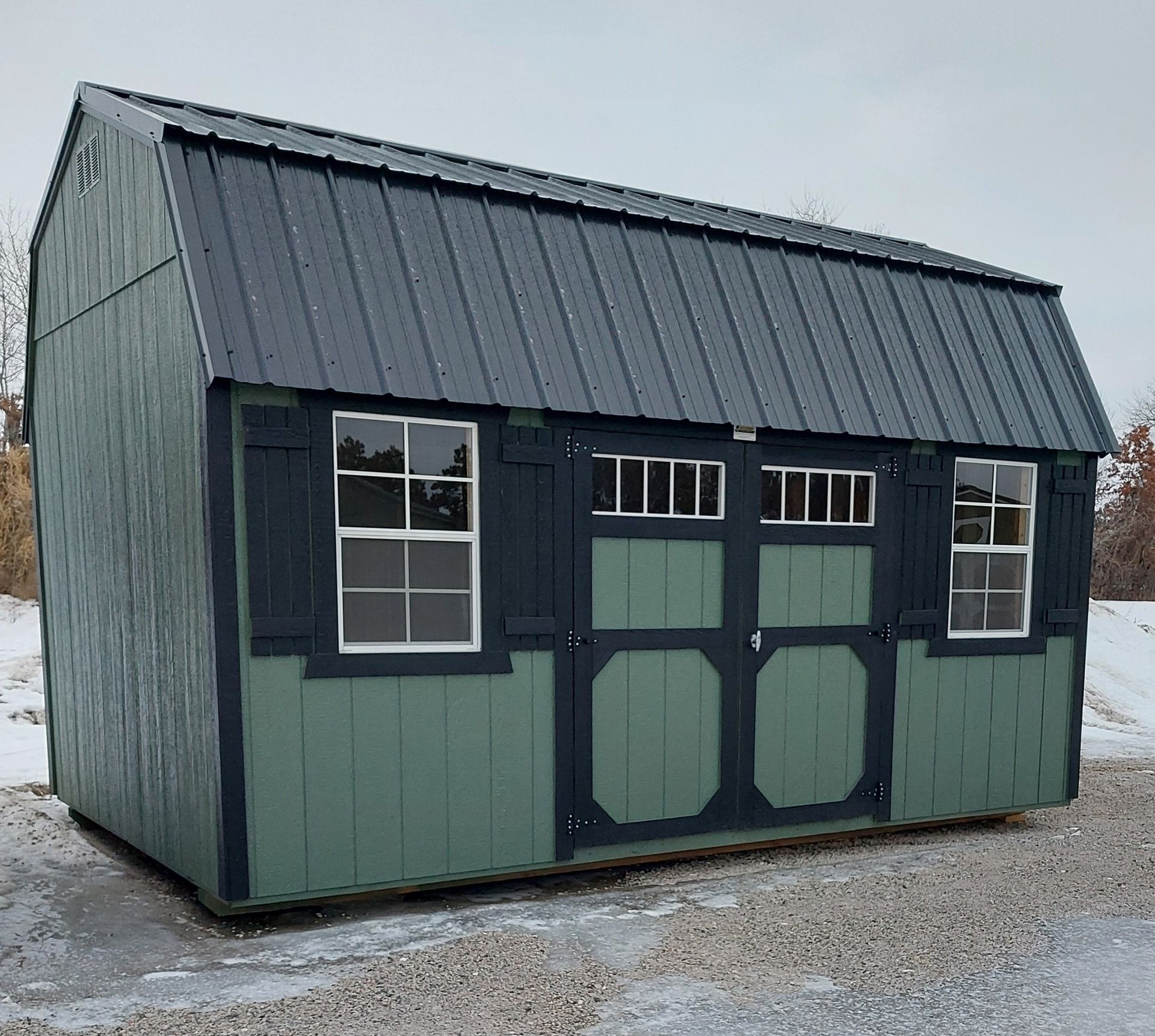 Sage green gambrel shed with black trim, a dark metal roof, two windows with shutters, and double doors on a gravel lot.