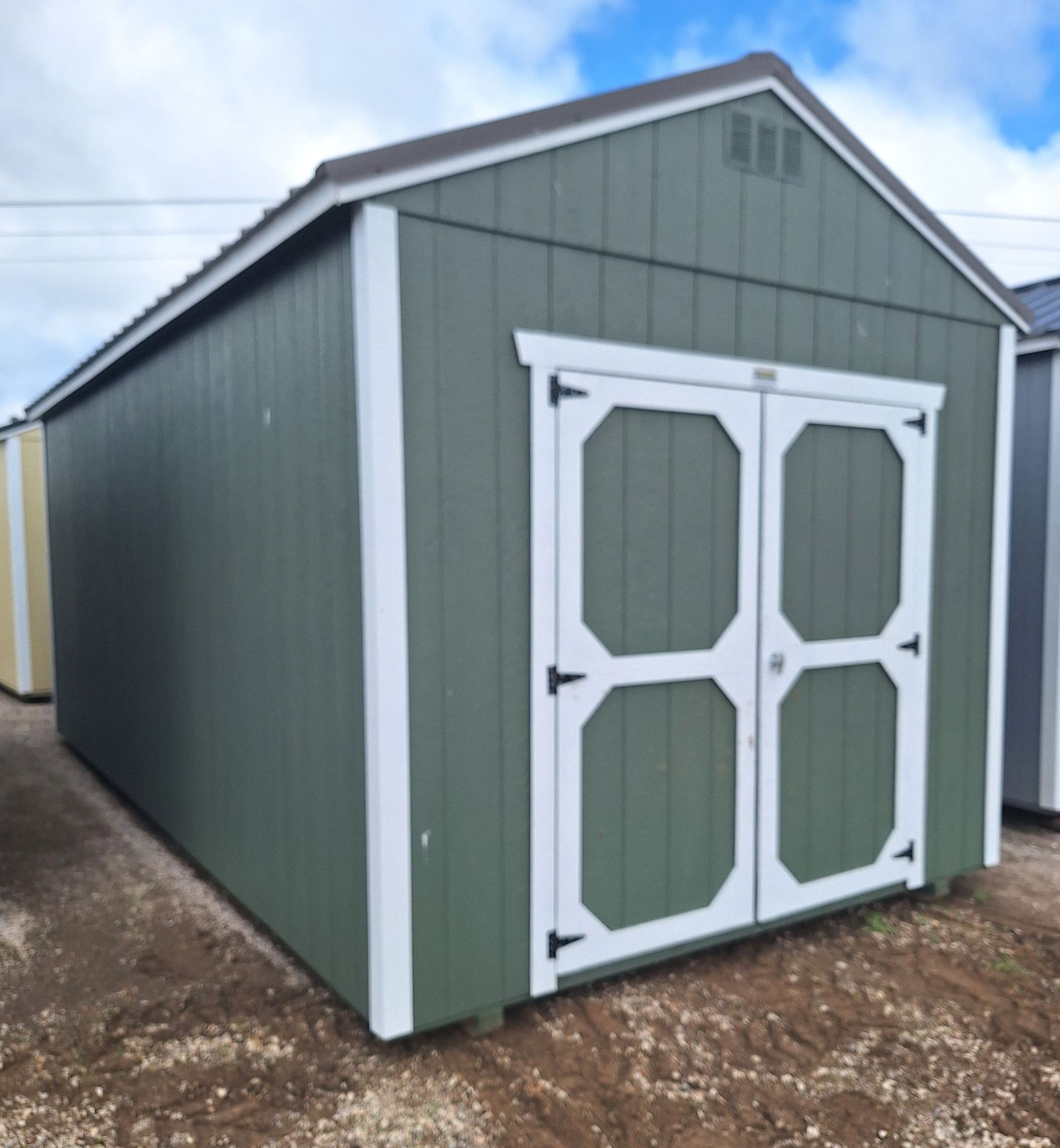 A green storage shed with a white-trimmed double door, angled outdoors on a gravel lot under a cloudy sky.