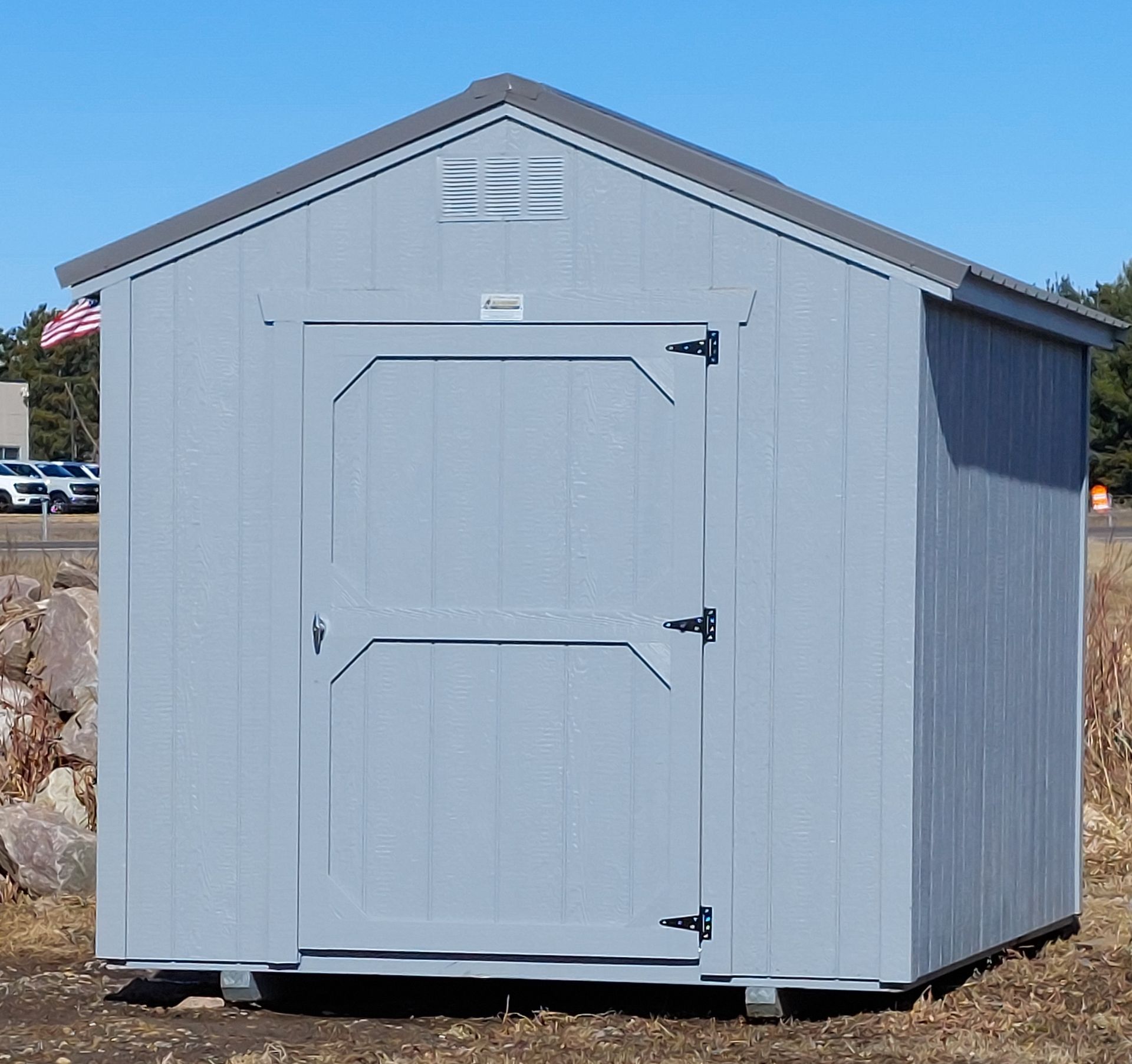 A light blue, wooden storage shed with a single door, a gable roof, and a vent, sitting on a gravel lot.