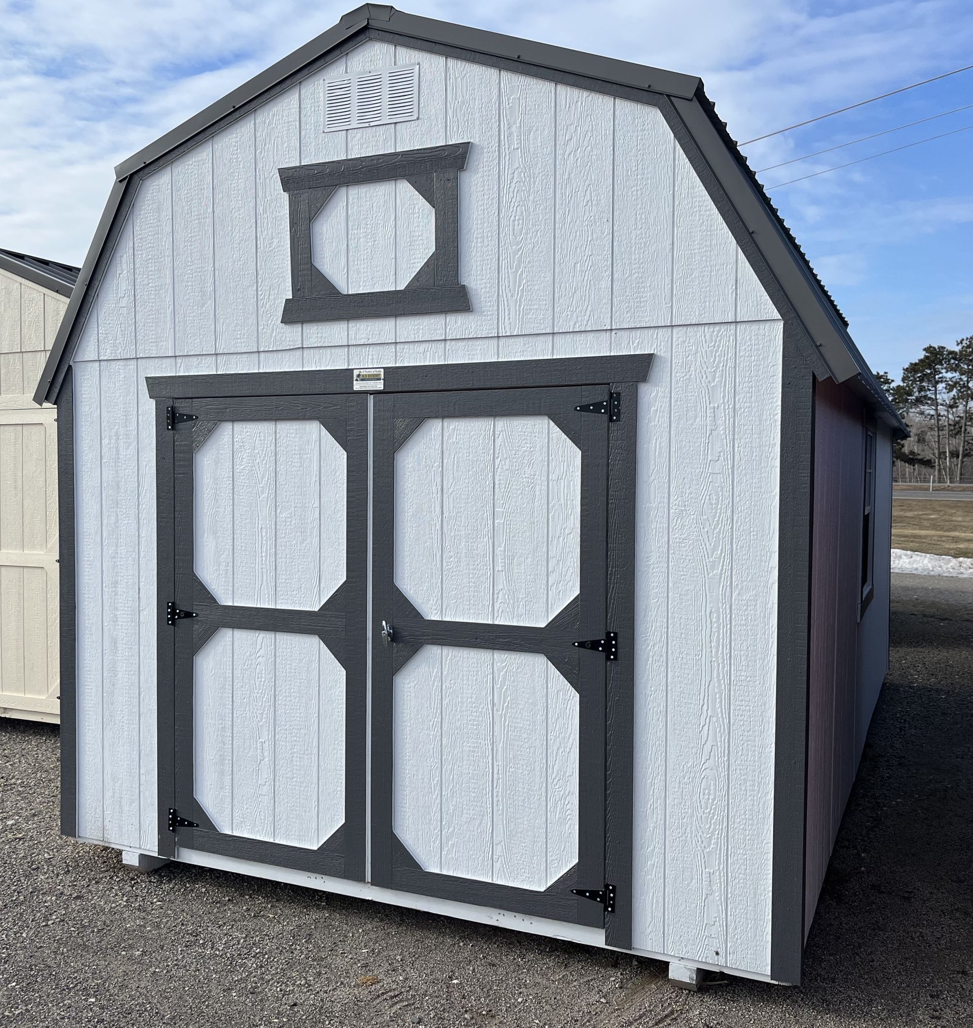 A light gray wooden shed with a gambrel roof and dark gray trim, featuring double doors and a decorative window above.