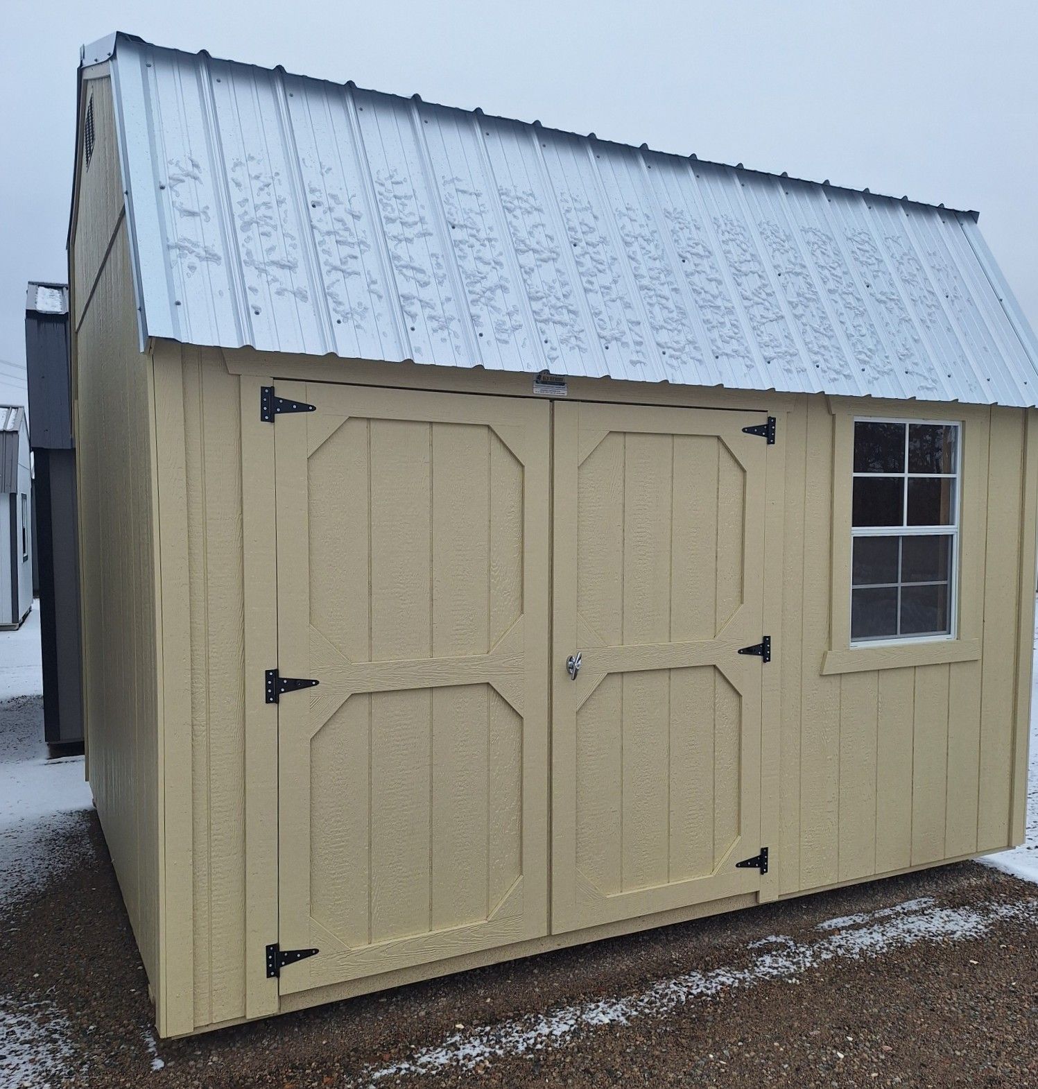 A yellow wooden storage shed with a silver metal roof, double doors, and one window, sitting on gravel in the snow.