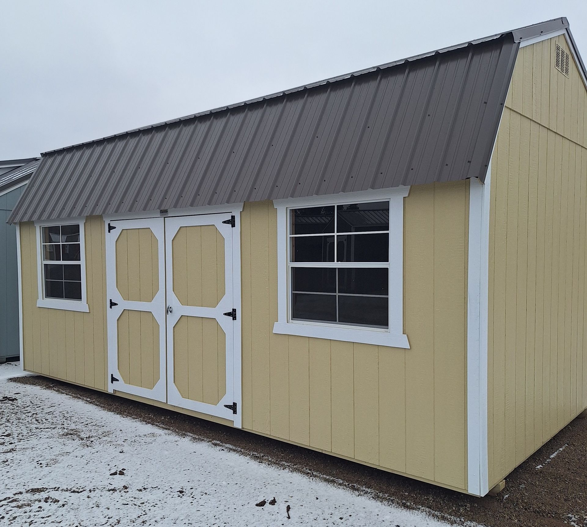 A yellow, single-story storage shed with a dark metal gambrel roof, white trim, double doors, and two windows.