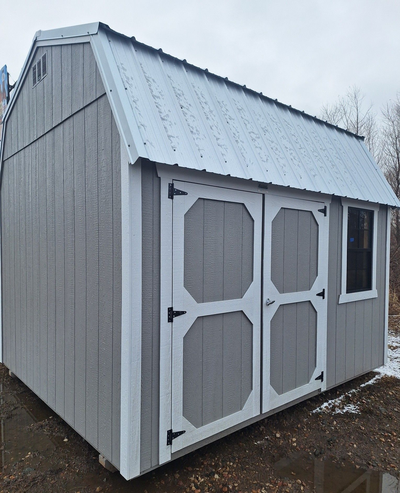 A gray wooden storage shed with a white trim, metal roof, double doors, a window and a workbench inside.