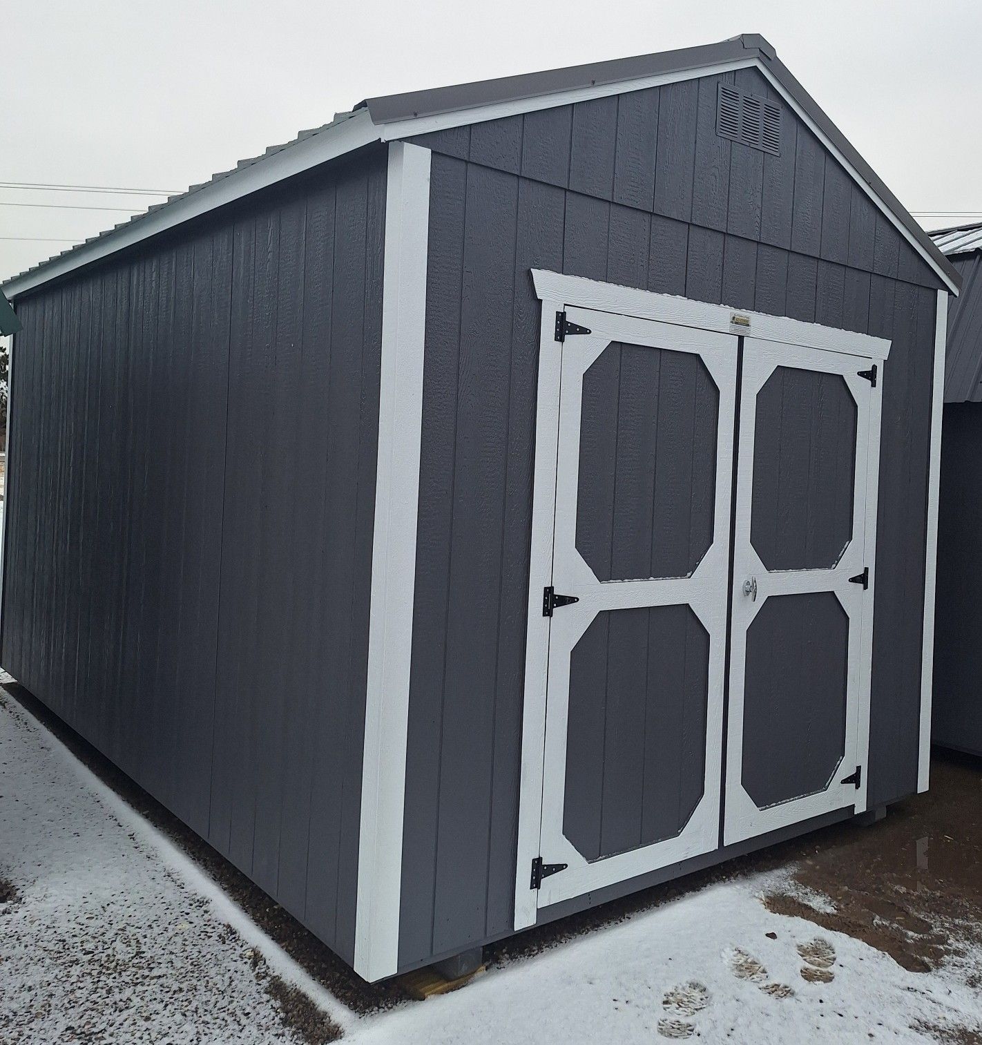 A dark gray wooden storage shed with white trim and double doors, standing on snowy ground under a cloudy sky.