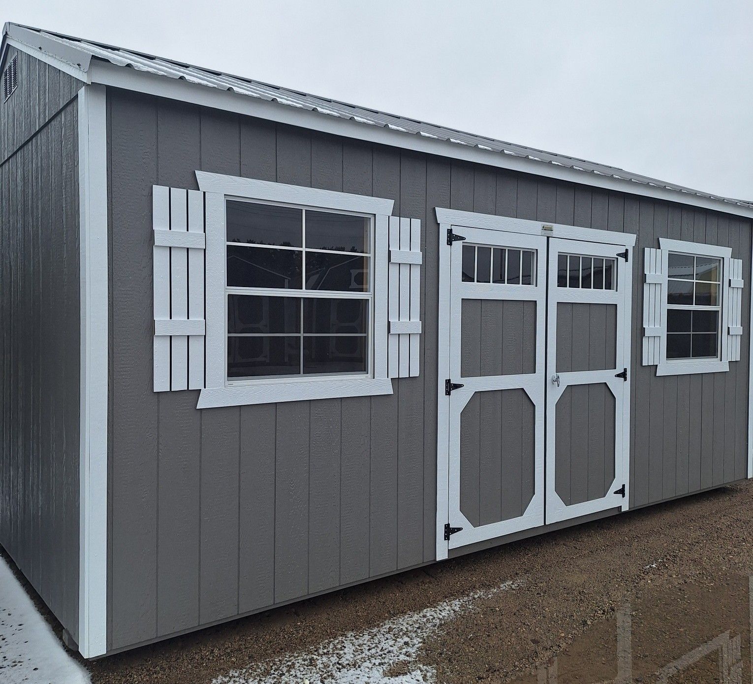 A gray storage shed with white trim, a double door, two windows with shutters, and a metal roof on a gravel surface.