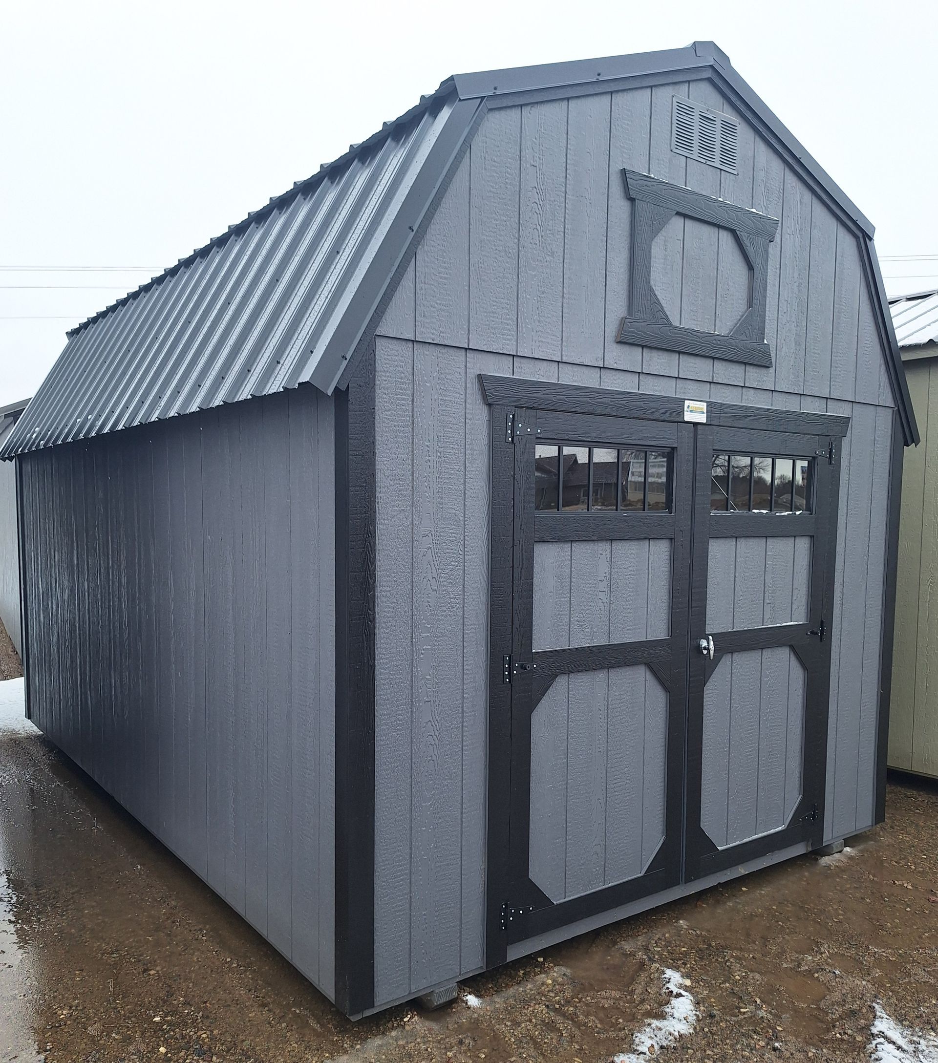 A gray wooden barn-style storage shed with a black metal roof, trim, and double doors with windows, standing on dirt.