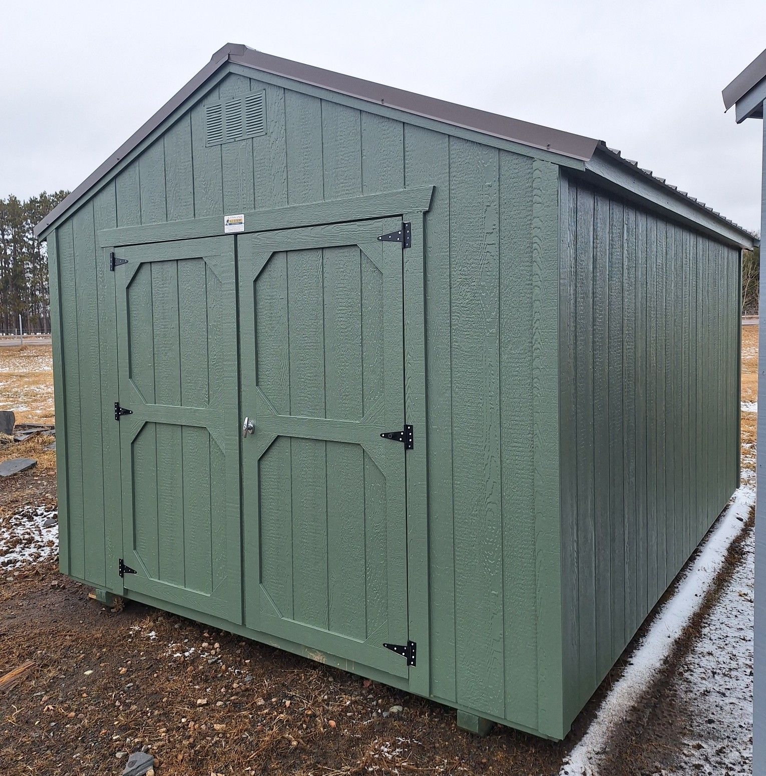 A green wooden storage shed with double doors, set on a gravel surface outdoors under a cloudy sky.