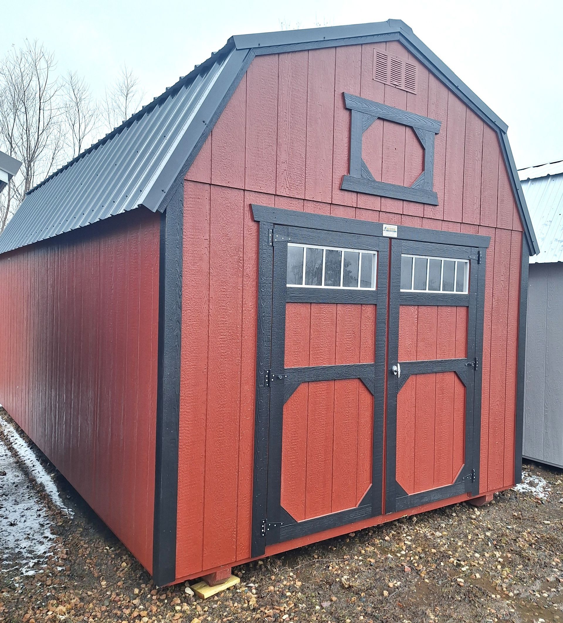 A red gambrel-roof shed with black trim and double doors, standing on a gravel lot with patches of snow.