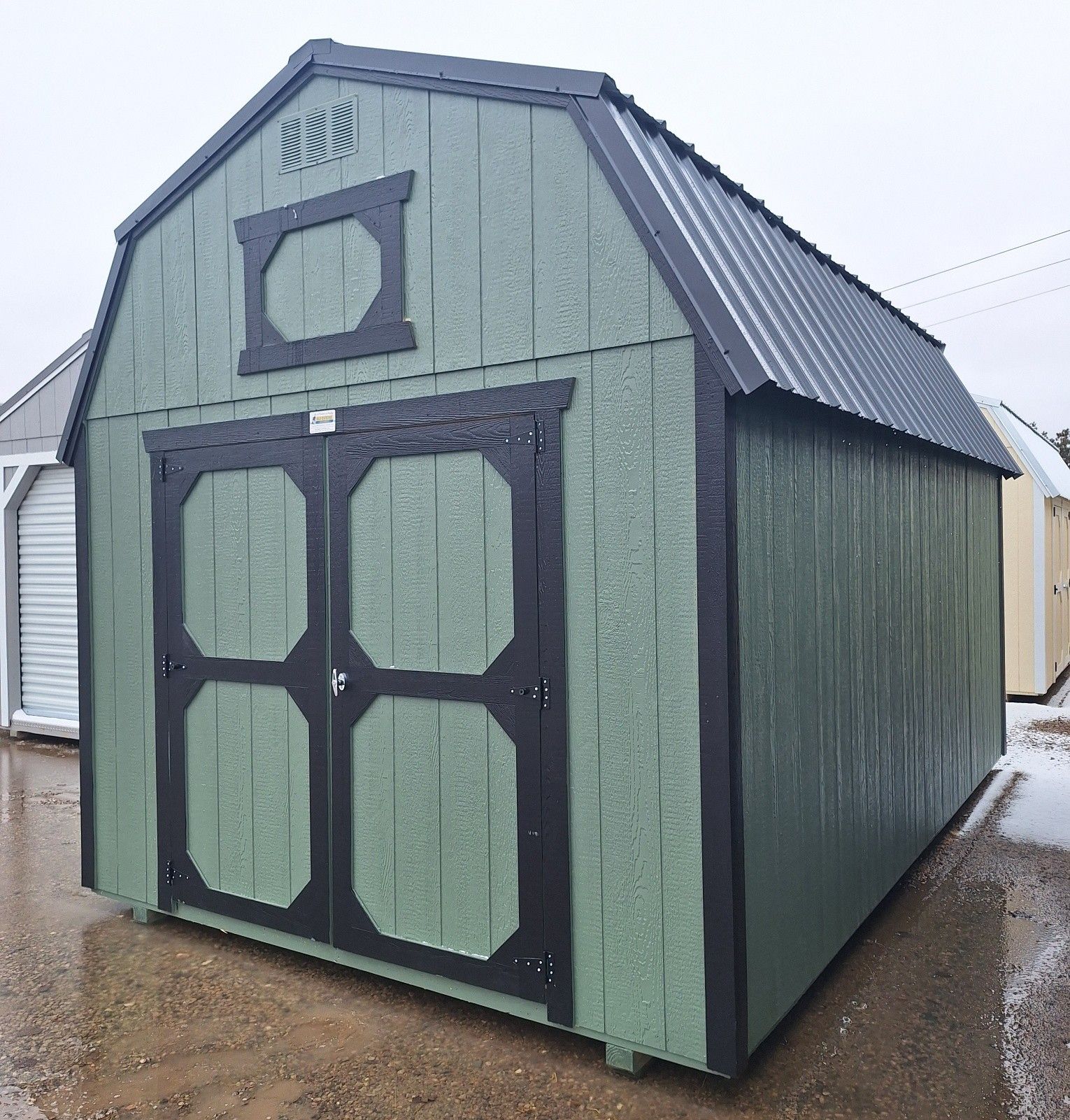 Sage green barn-style shed with black trim and a metal roof, shown on a gravel lot during winter.