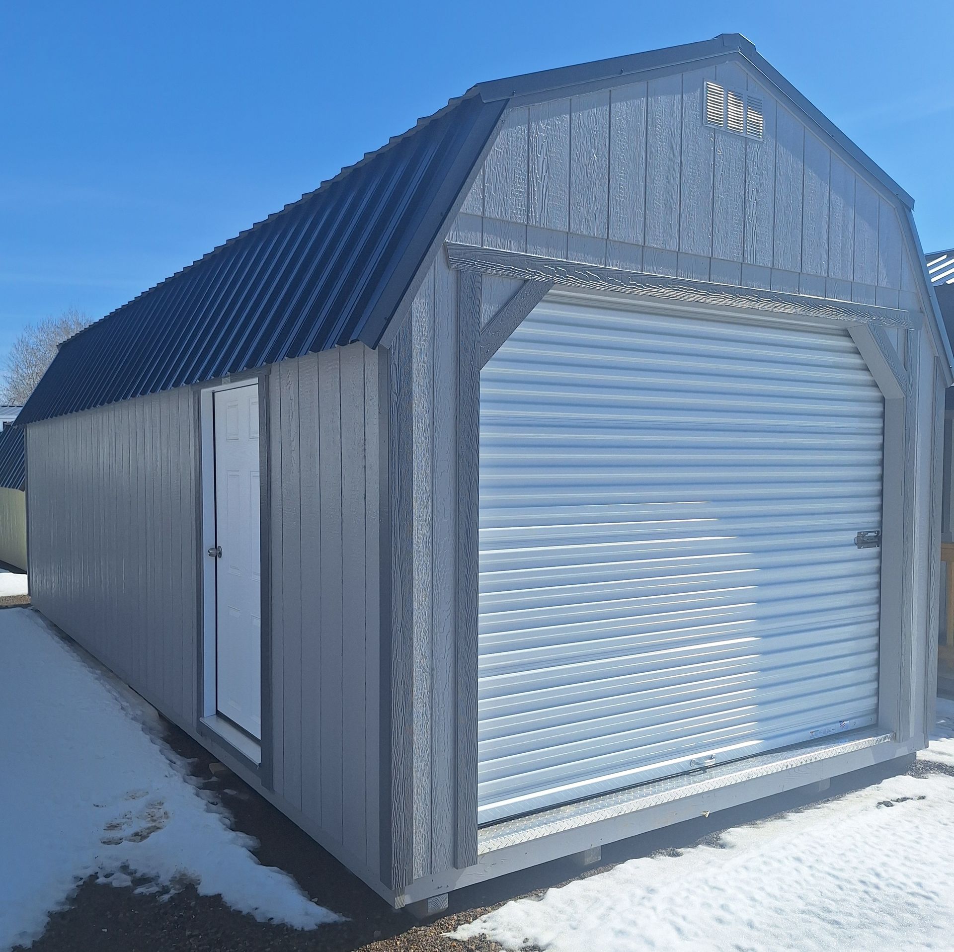 A gray storage shed with a dark metal gambrel roof, a side entry door, and a white roll-up garage door on a snowy lot.