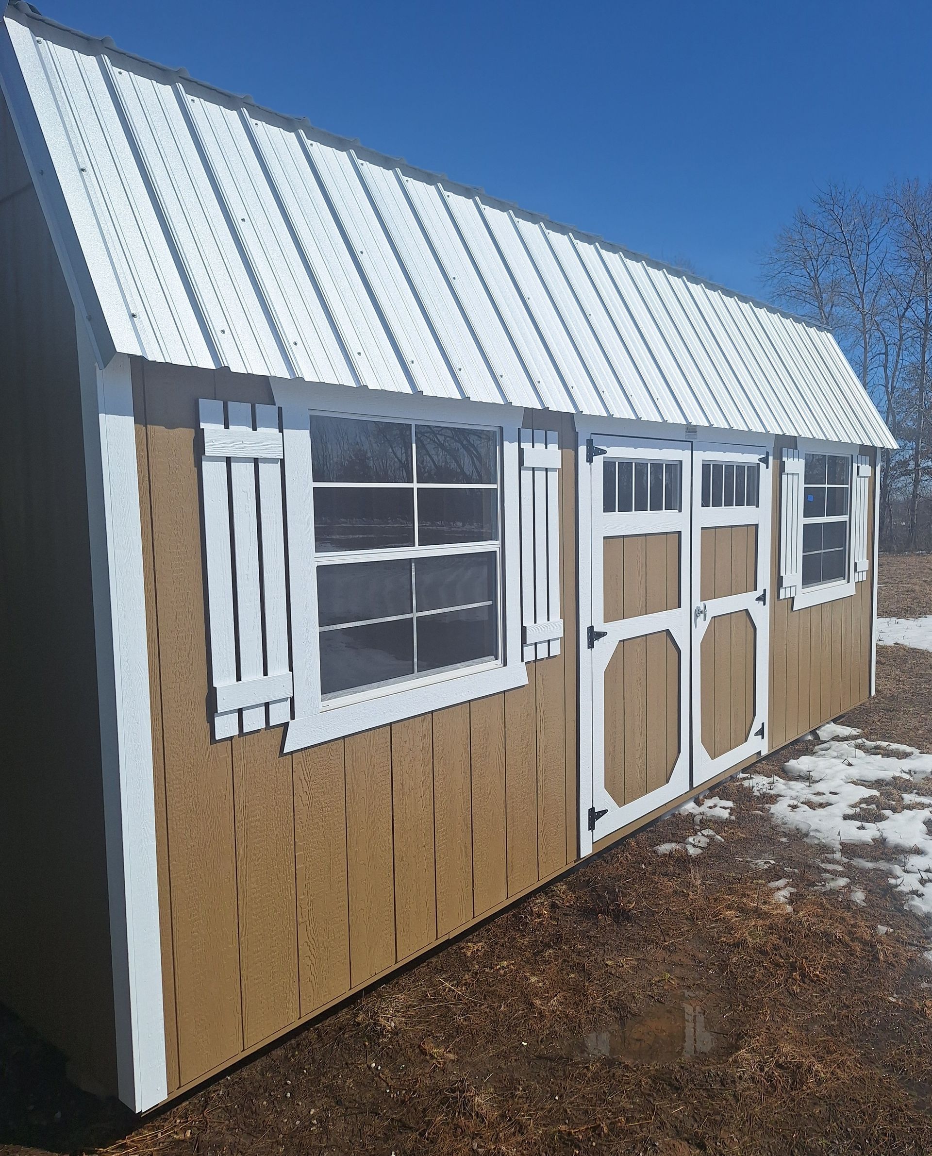Tan shed with a white metal roof, white trim, shutters, a window, and double doors, situated on snowy ground.