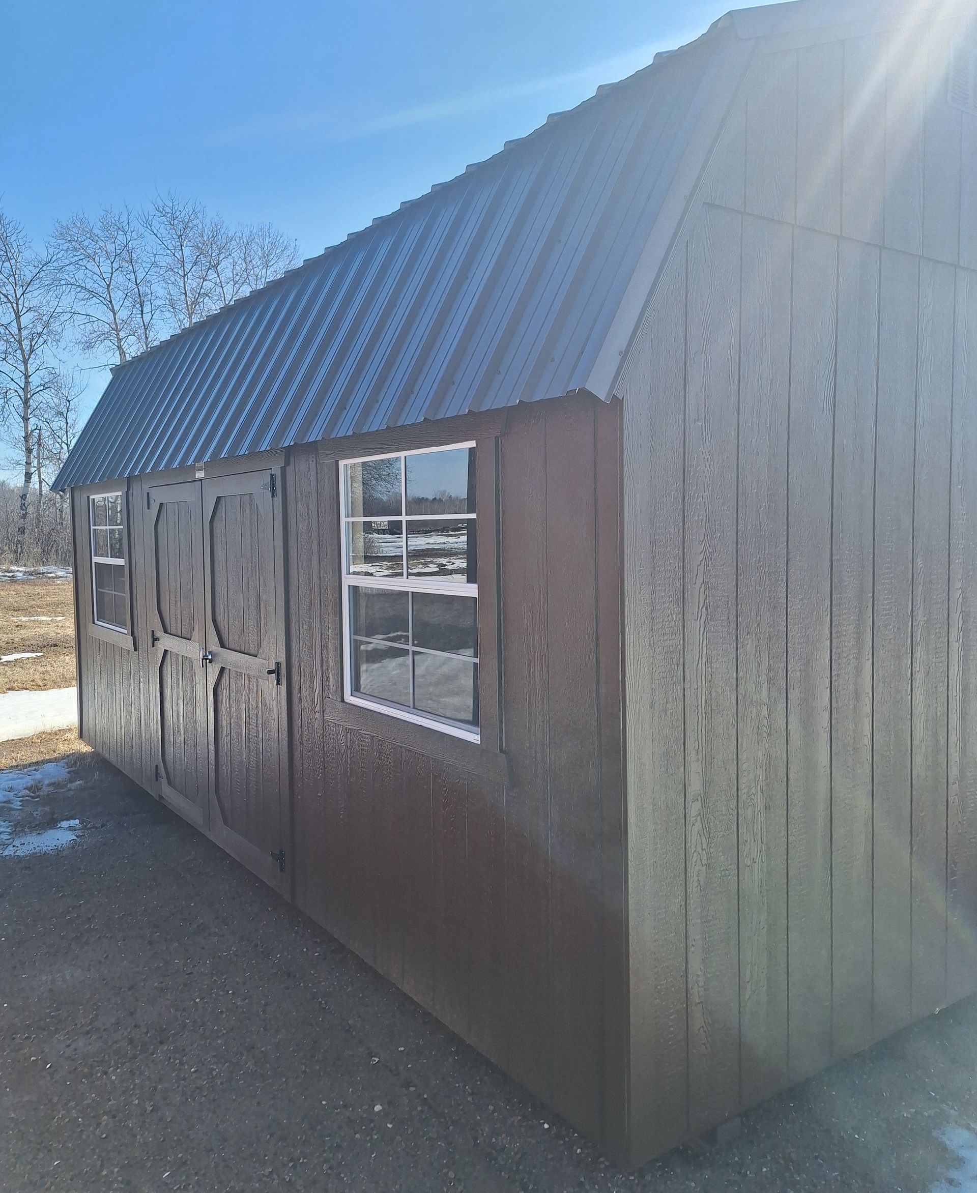 A brown wooden shed with a metal roof and two windows, set in a gravel area under a clear blue sky.