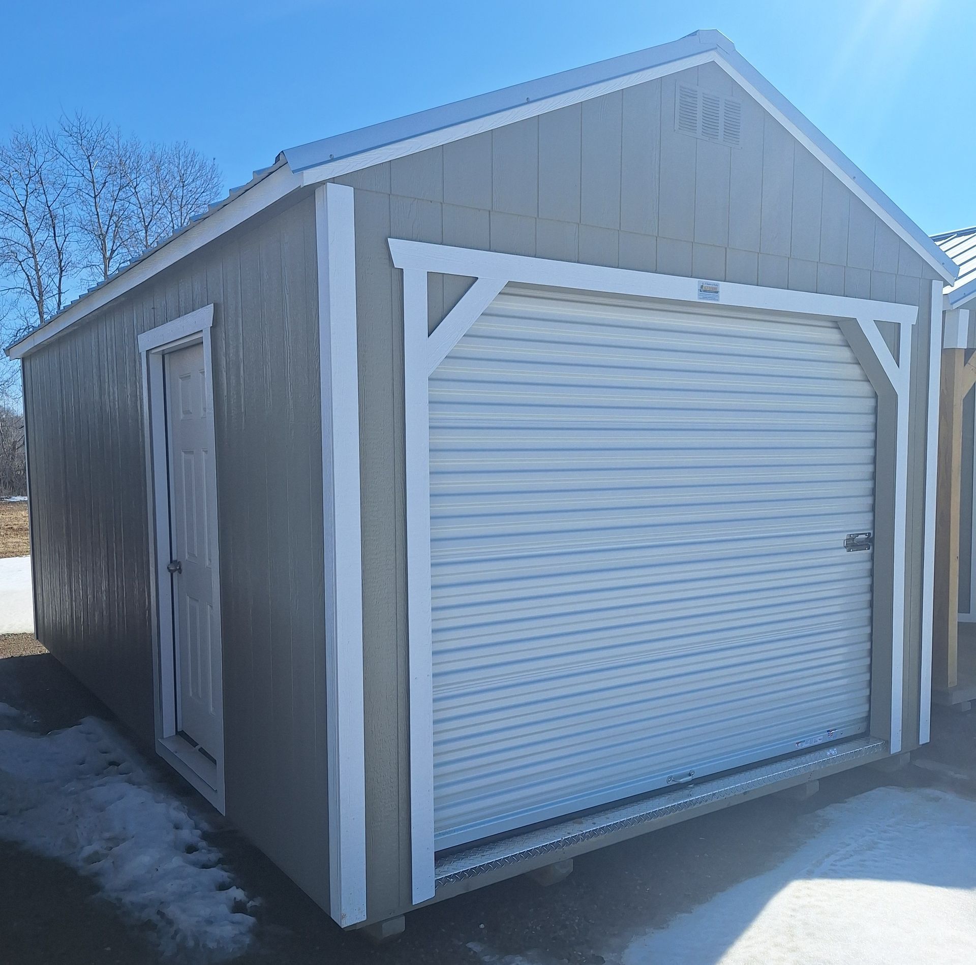 A gray, rectangular storage shed with a white trim, a roll-up garage door, and a side access door, set on a snowy surface.