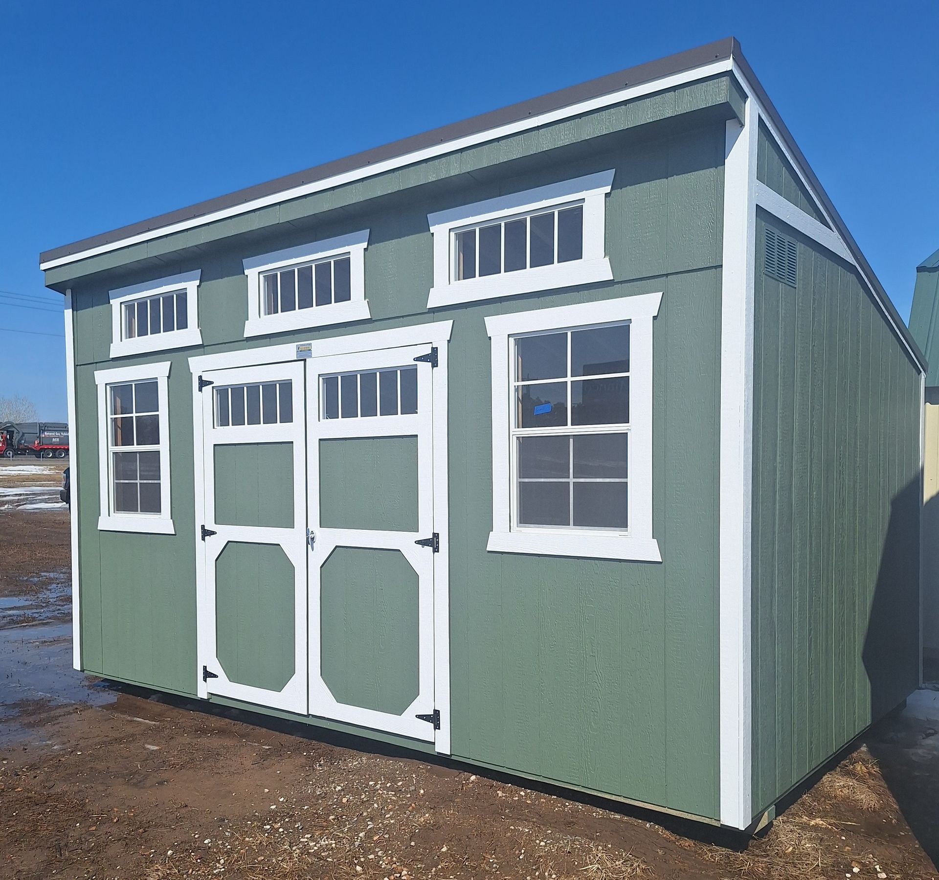Sage green wooden shed with a slanted roof, white trim, double doors, two standard windows, and three transom windows.
