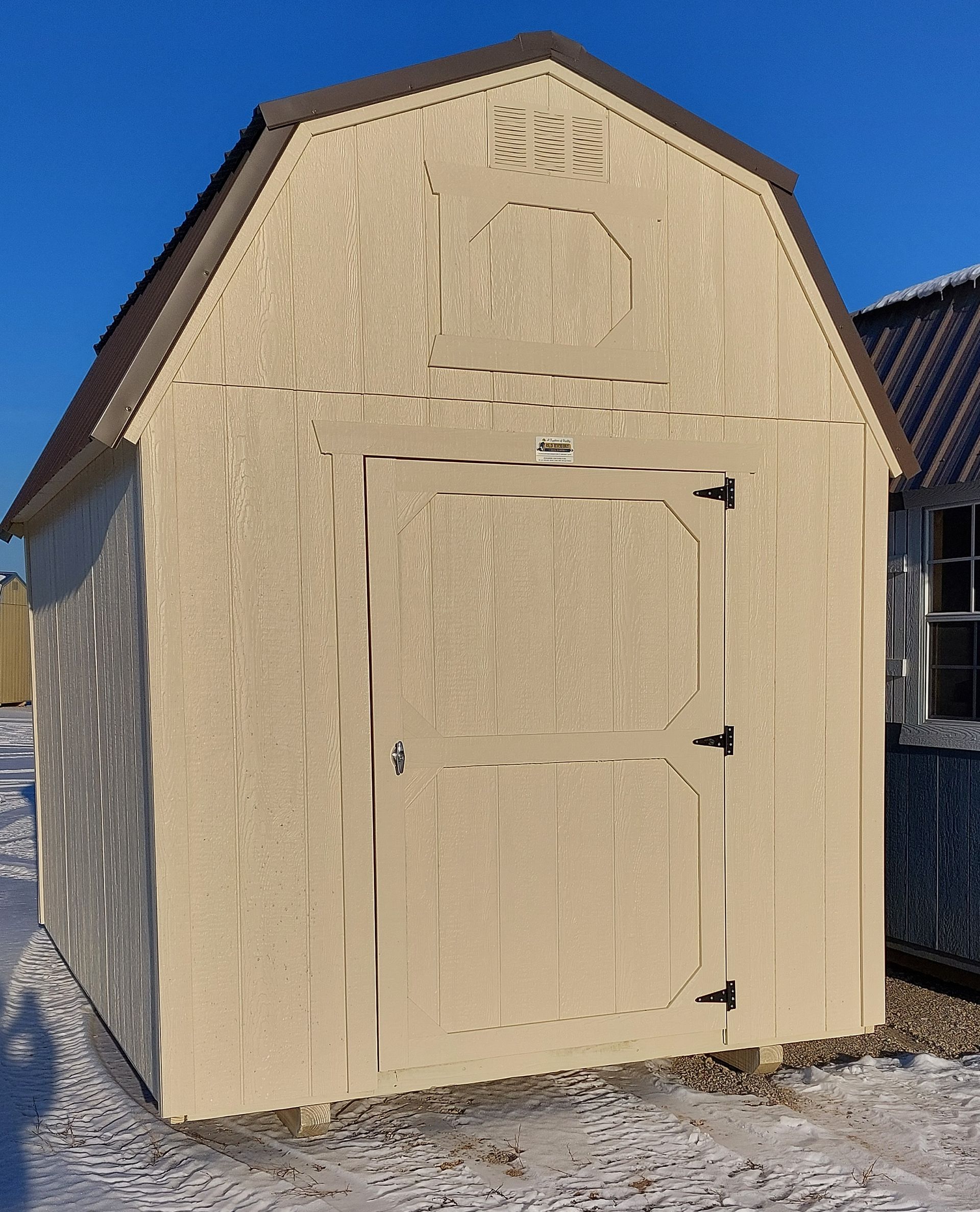 A tan-colored, gambrel-roof wooden storage shed with a single door, standing on snowy ground under a clear blue sky.