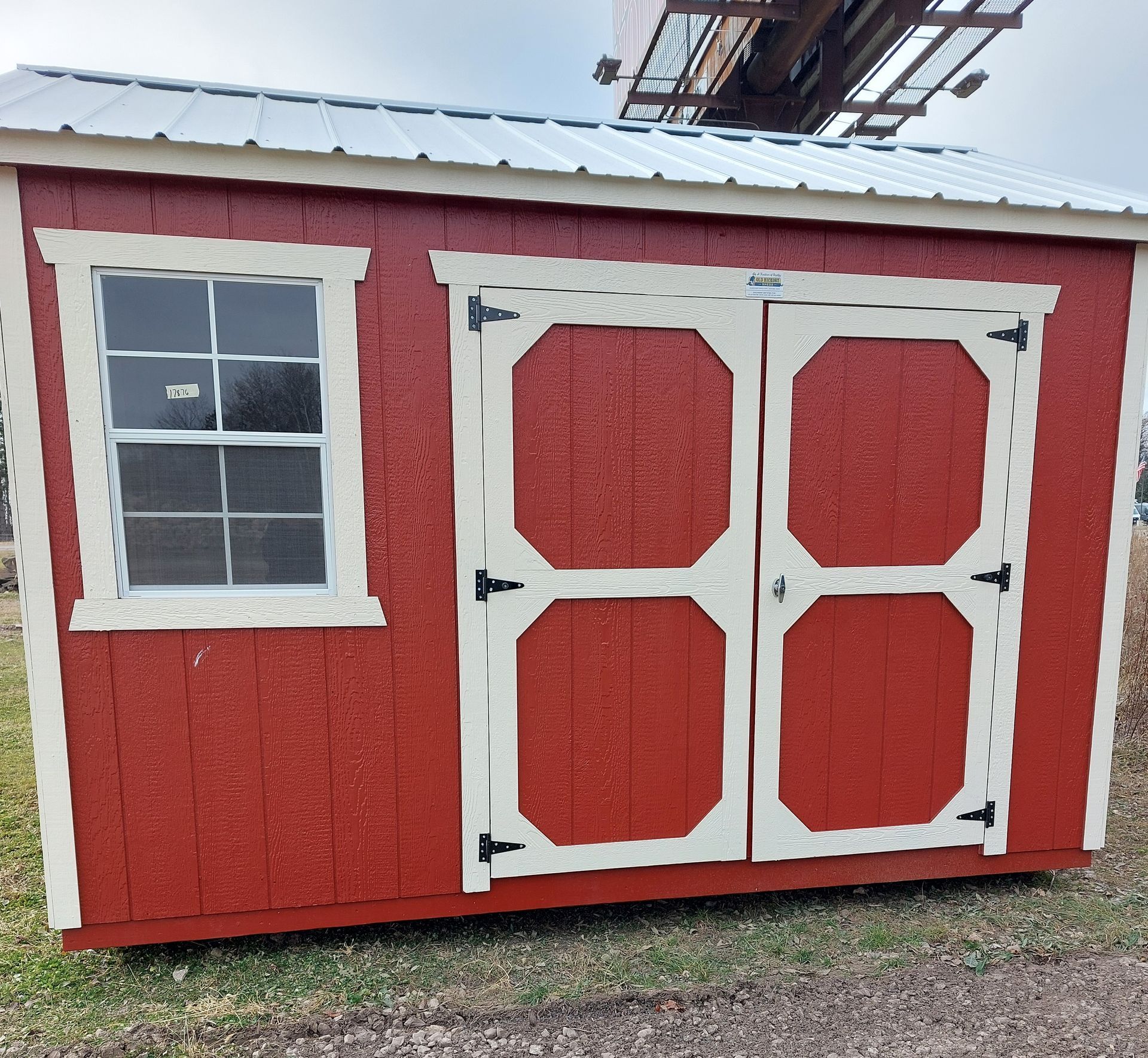 A red wooden storage shed with cream trim, a single window, and double doors, standing on a grassy outdoor lot.