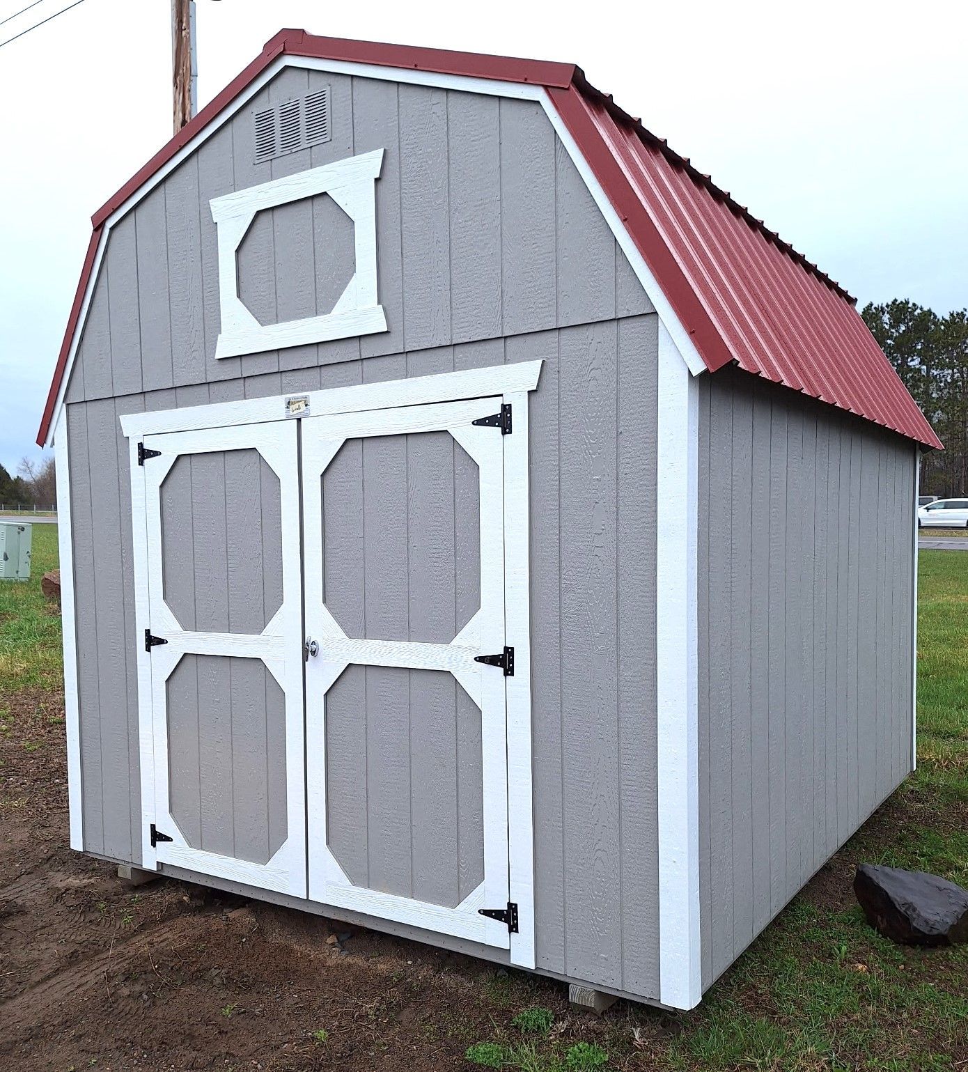 A gray wooden barn-style storage shed with white trim and a red metal roof, standing outdoors on a grassy lot.