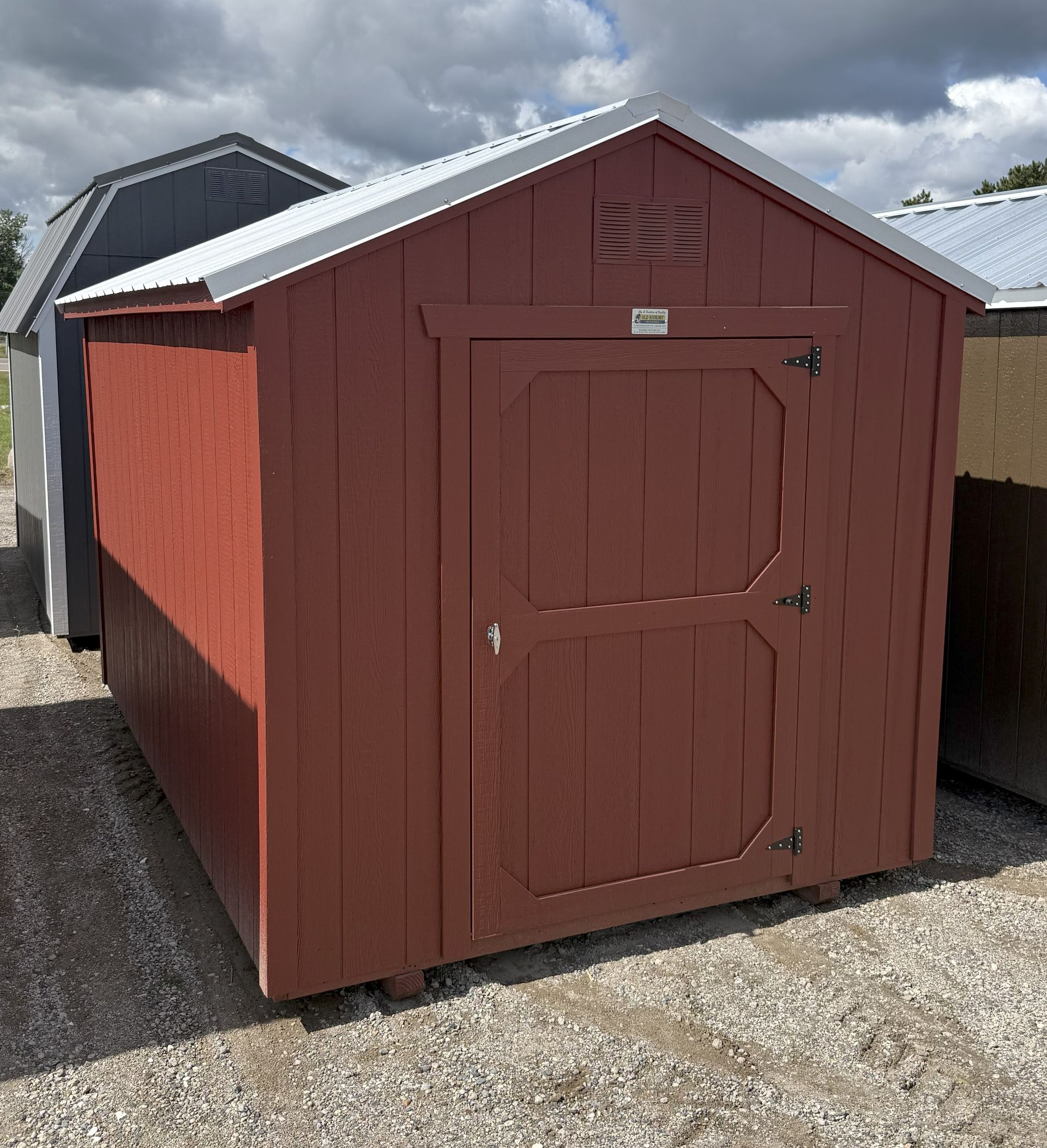 A red wooden storage shed with a silver metal roof, situated on a gravel surface outdoors under a cloudy sky.
