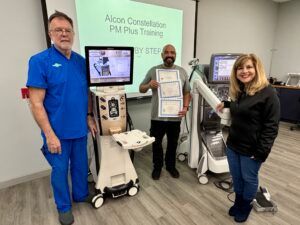 Three people stand with ophthalmic equipment, one holding certificates in a training room.