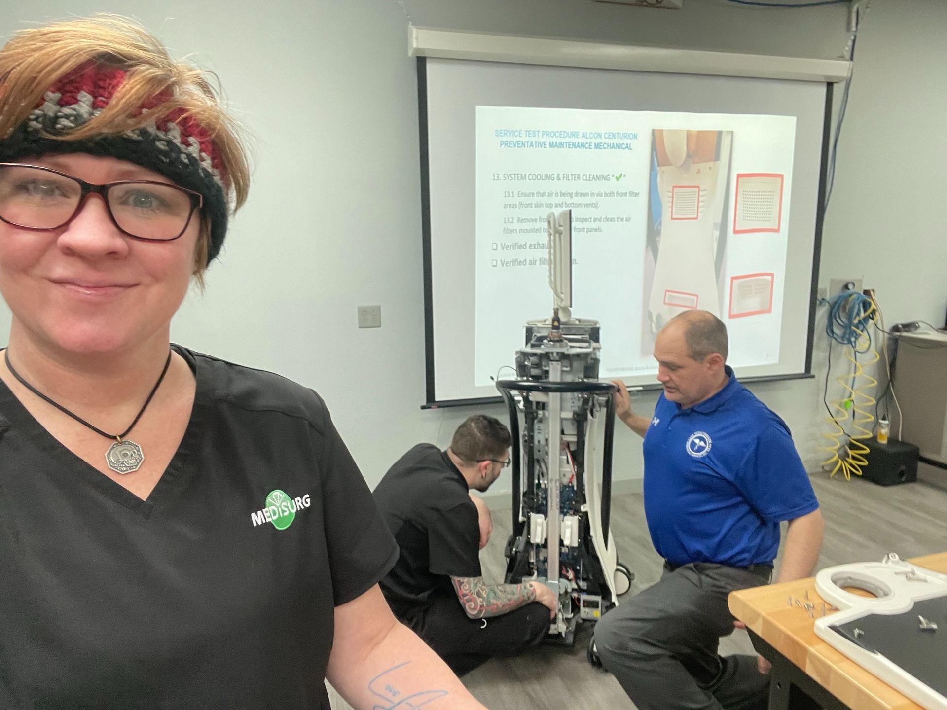 Woman in glasses smiles at the camera, two men work on equipment with a presentation on a screen behind them.