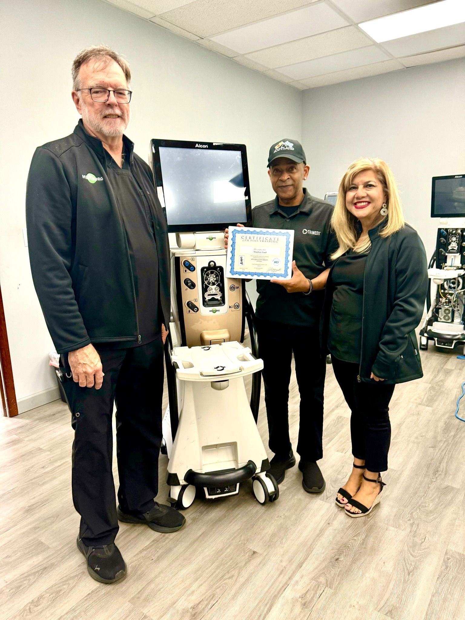 Three people stand by medical equipment, holding a certificate. Indoors, with grey and white decor.