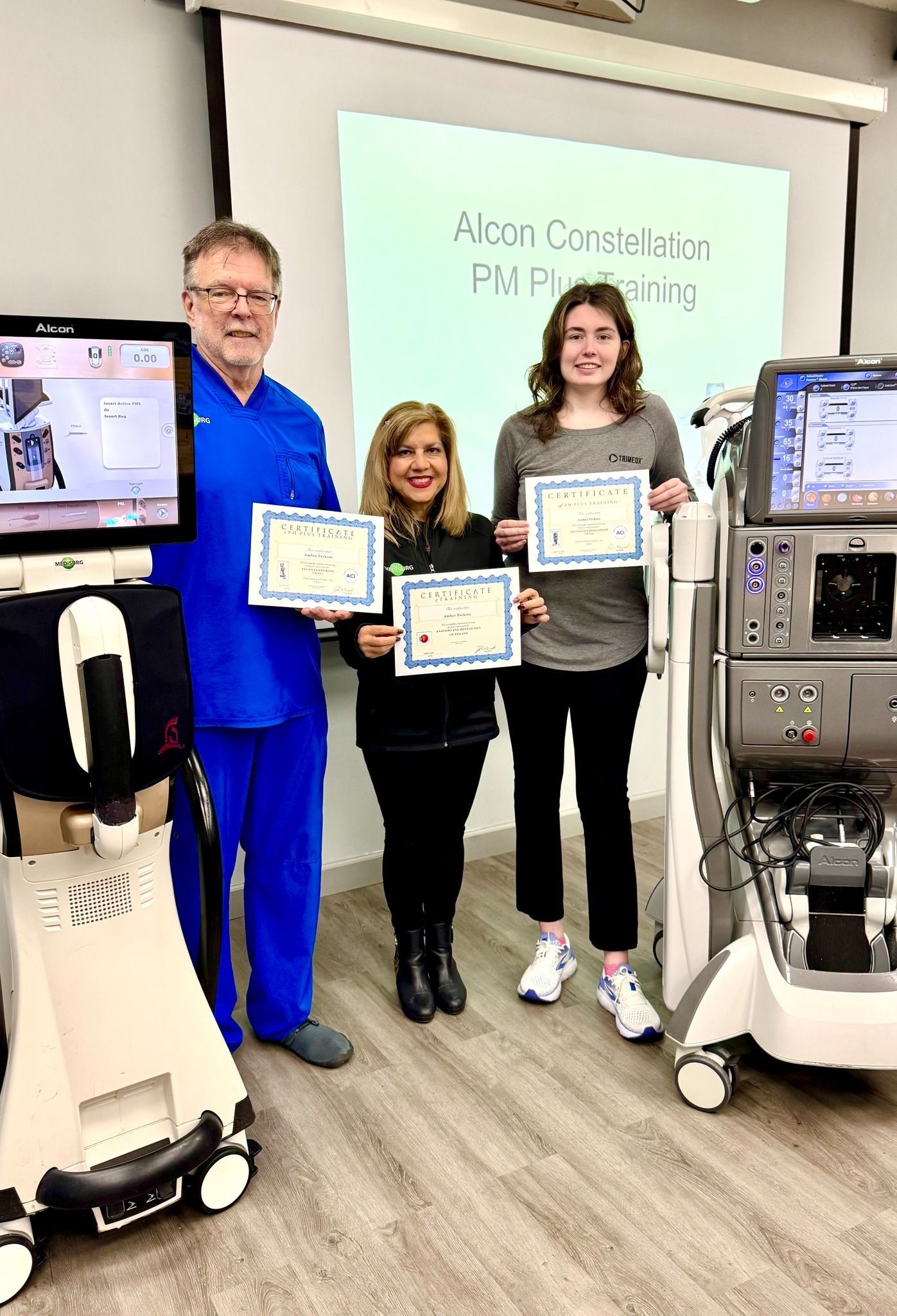 Three people holding certificates, standing in front of medical equipment and a projector screen.