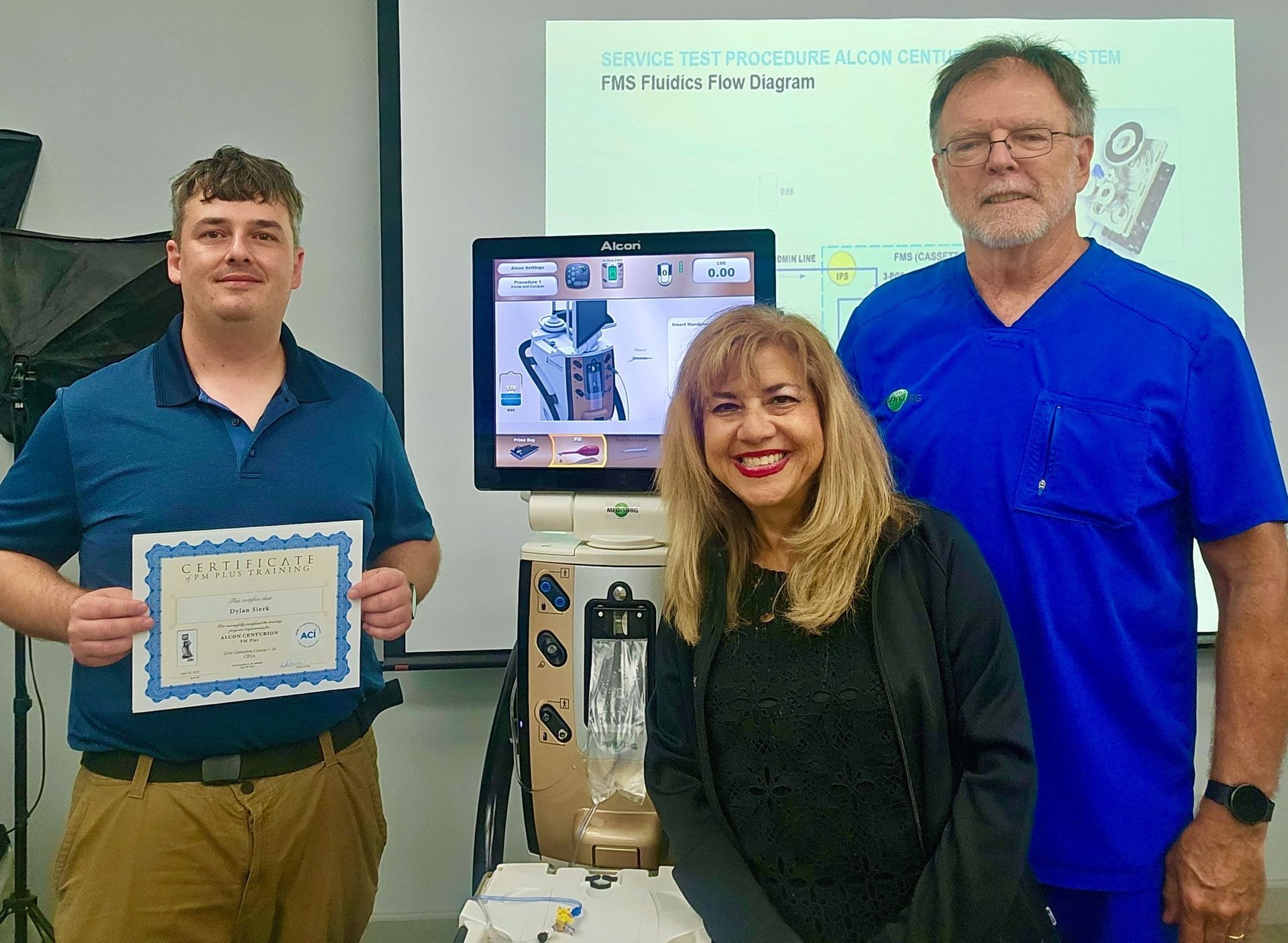 Three people pose with medical equipment; man holds certificate, woman smiles, man in scrubs stands.
