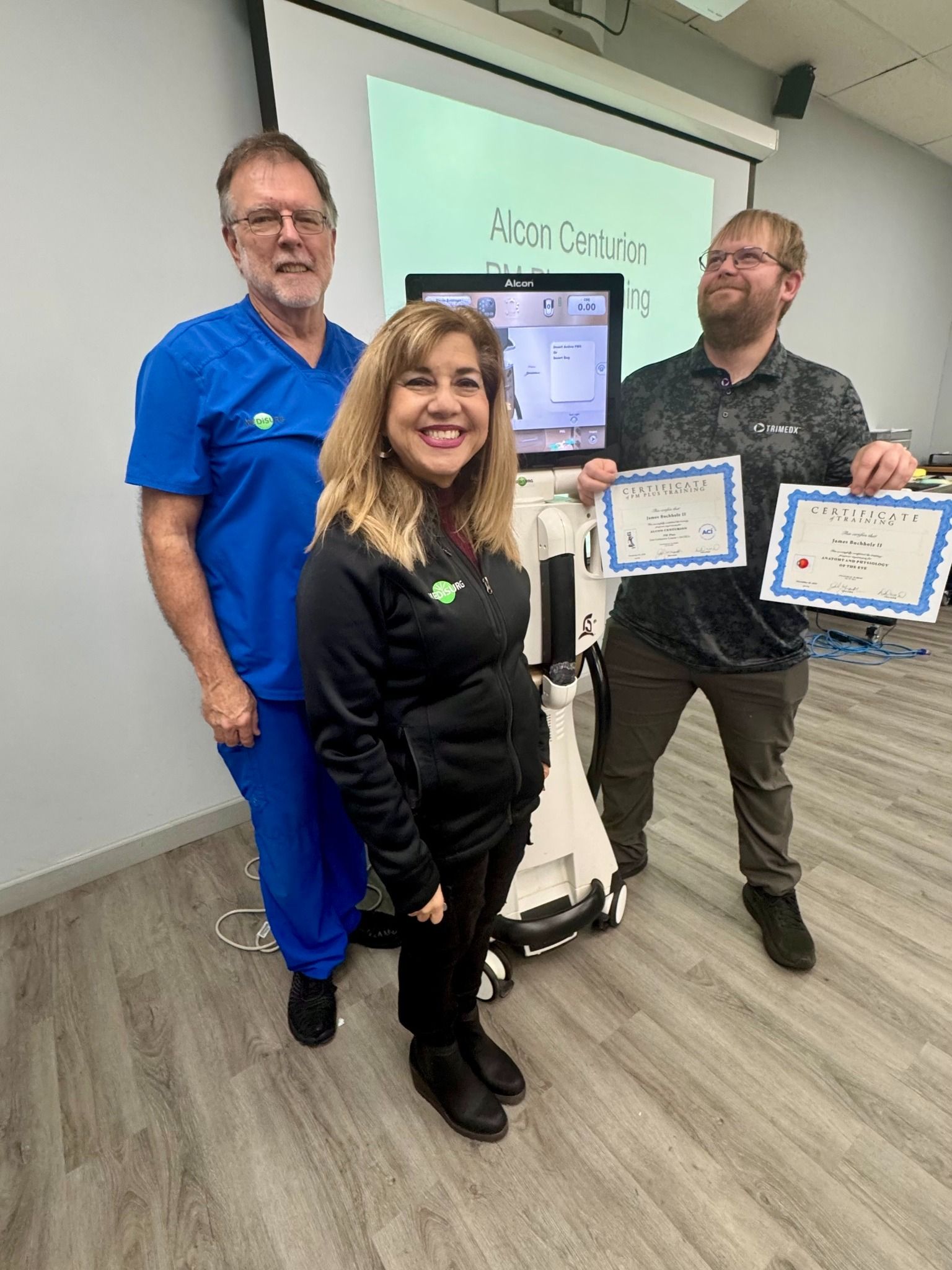 Three people stand with a medical device, holding certificates. Two smile, and one is in blue scrubs.