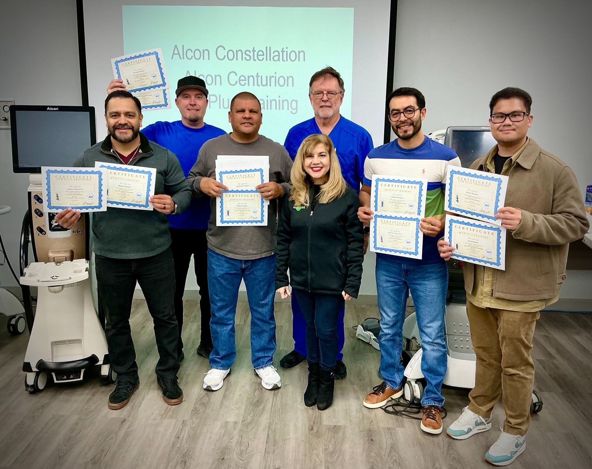 Group of people holding certificates, smiling, at a training session. Medical equipment is visible in the background.