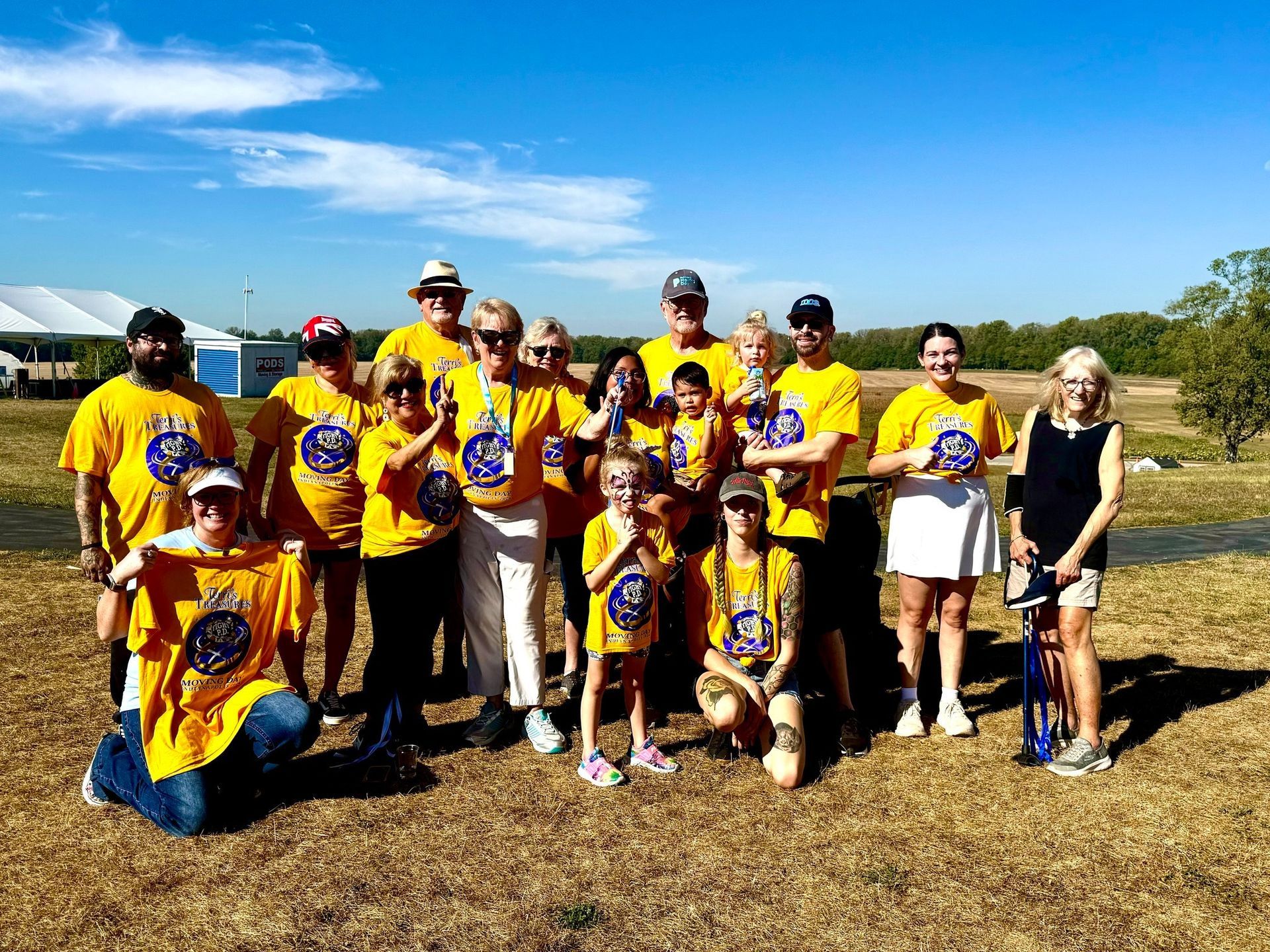 Group of people in yellow shirts, standing in a field under a blue sky. Some hold up shirts and pose.