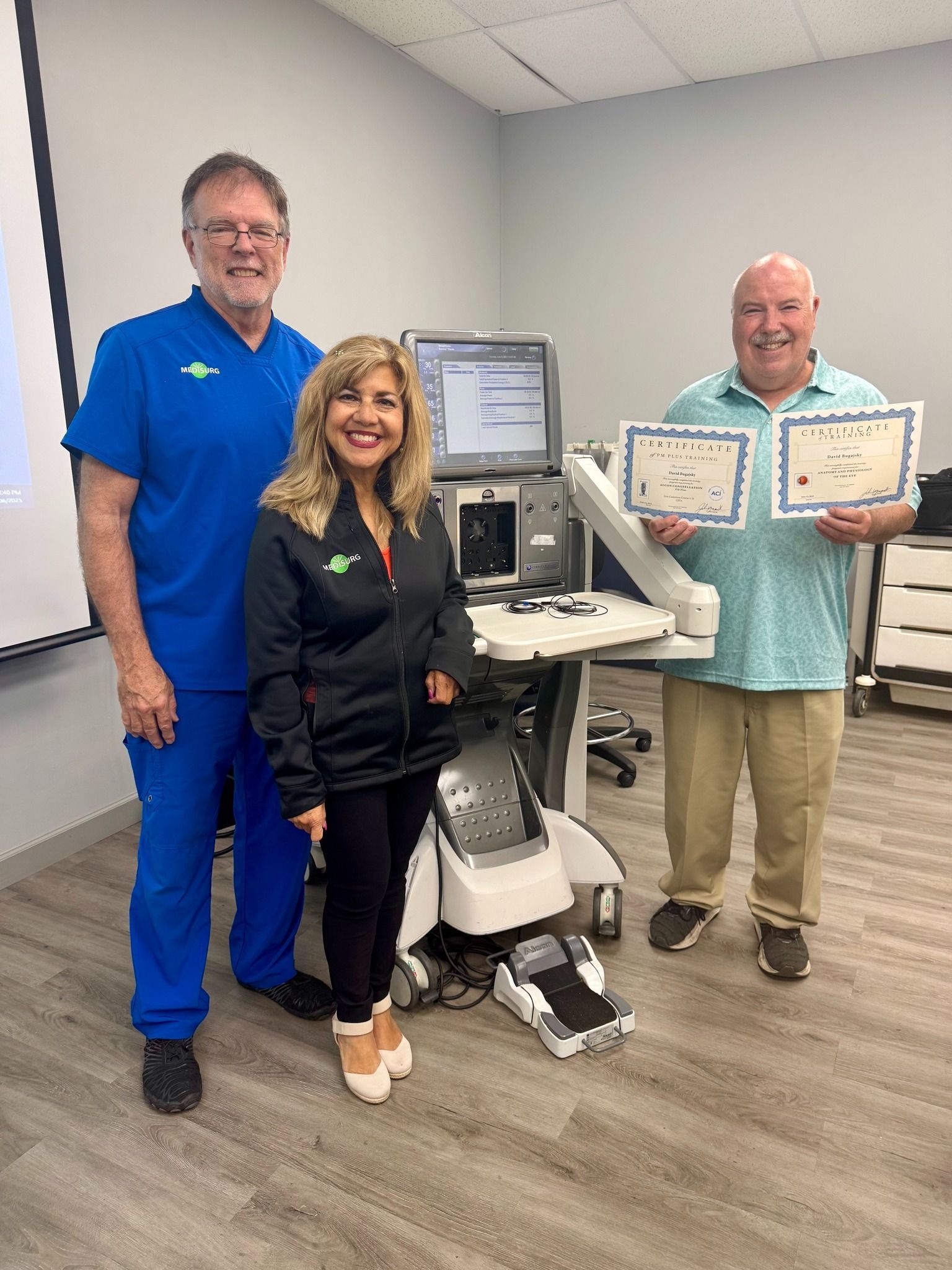 Three people stand next to medical equipment. A woman and a man hold certificates. The man on the left wears blue scrubs.