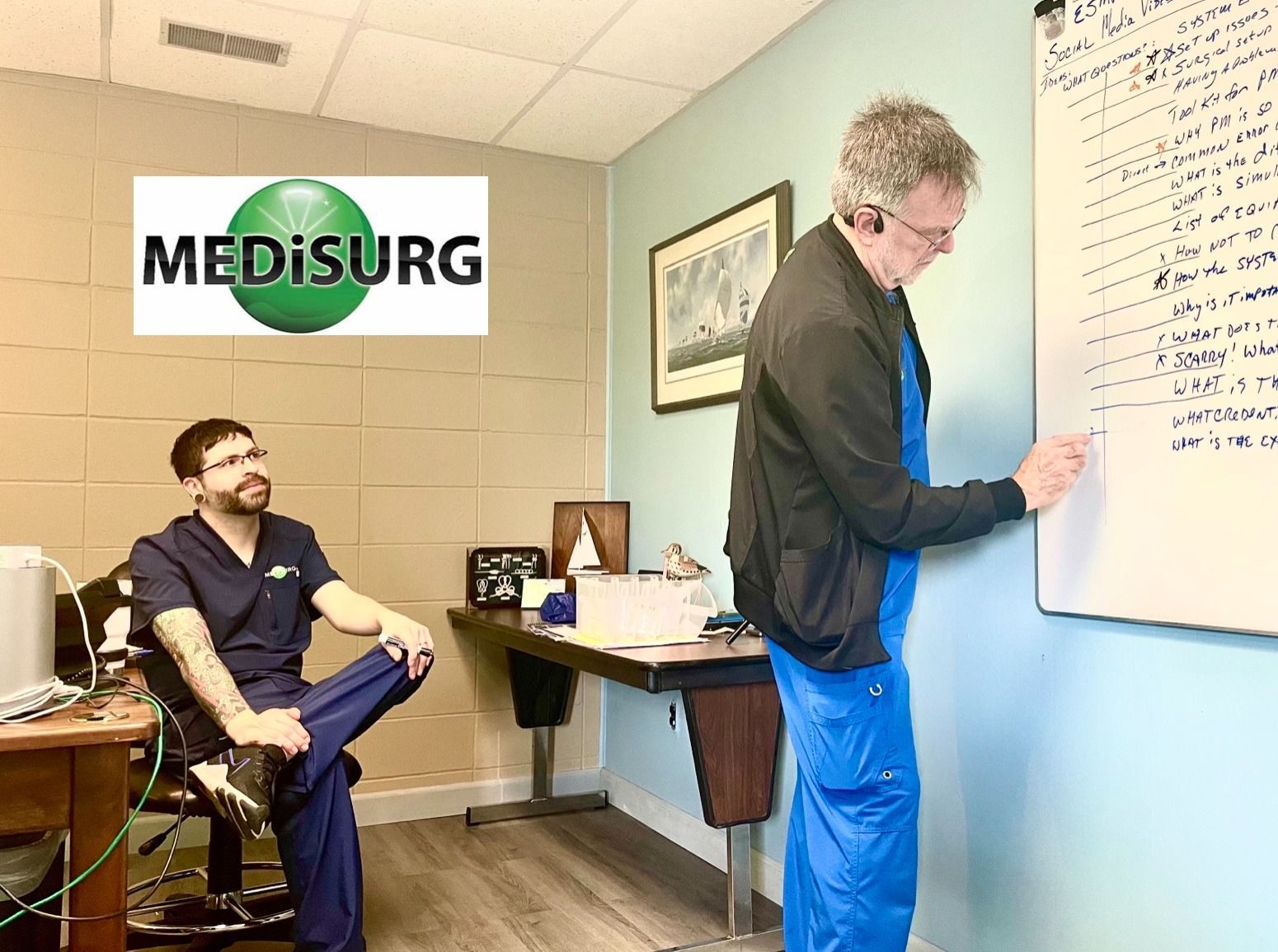 Two healthcare workers in an office. One writes on a whiteboard, the other sits and watches. 
