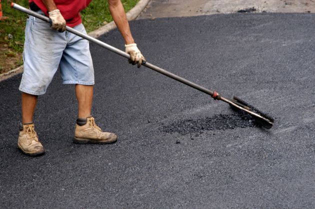 A worker operating a white road-marking machine while another person in a safety vest walks alongside on a paved road.