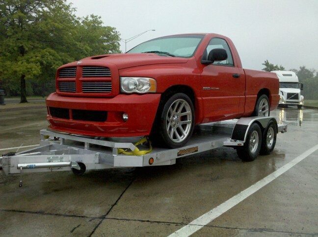 Red Dodge Ram truck on a silver trailer, parked on wet pavement near a white truck, under overcast skies.