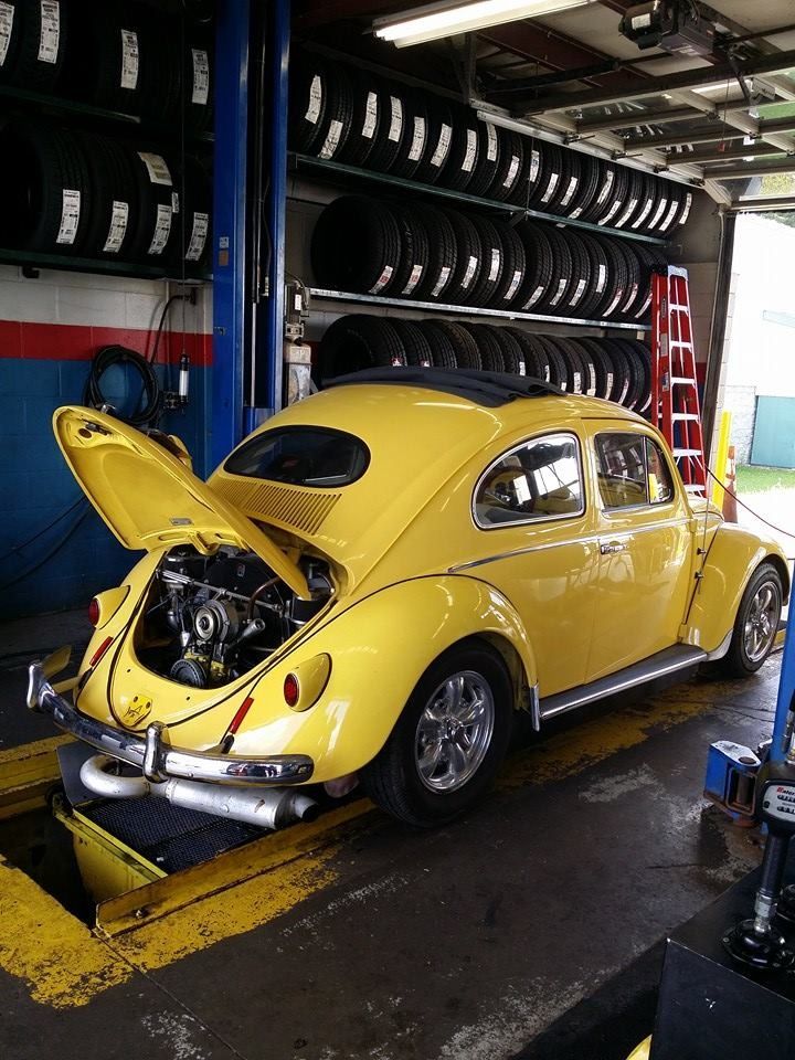 Yellow Volkswagen Beetle in a repair shop with the engine exposed, tires in the background.