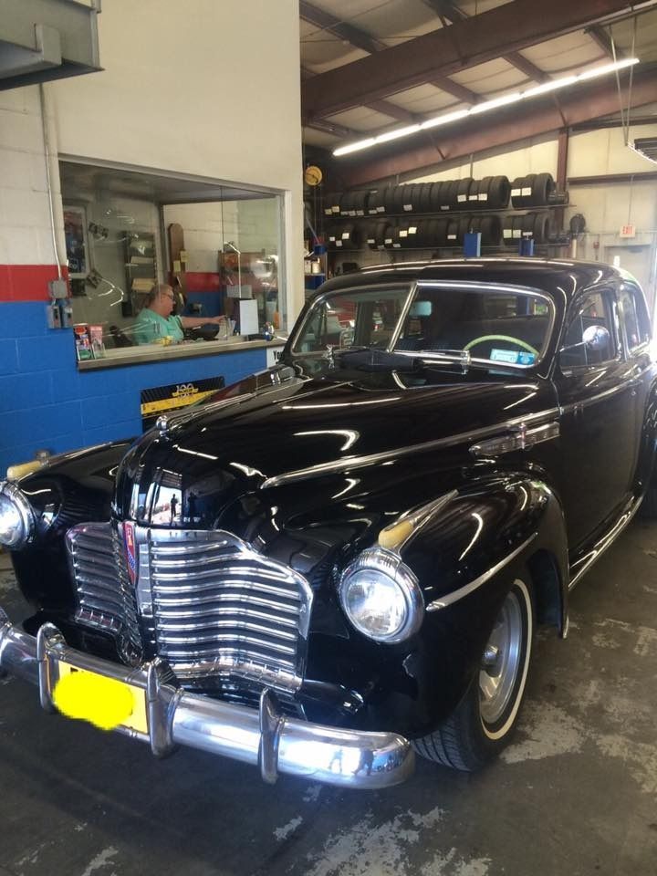 Black vintage Buick in a garage, shiny with chrome details. A person sits at a counter in the background.