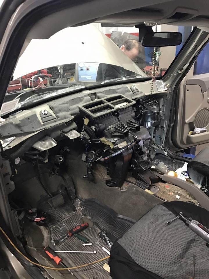 Interior of a car with dashboard dismantled. Tools and parts scattered; person visible in background with hood open.