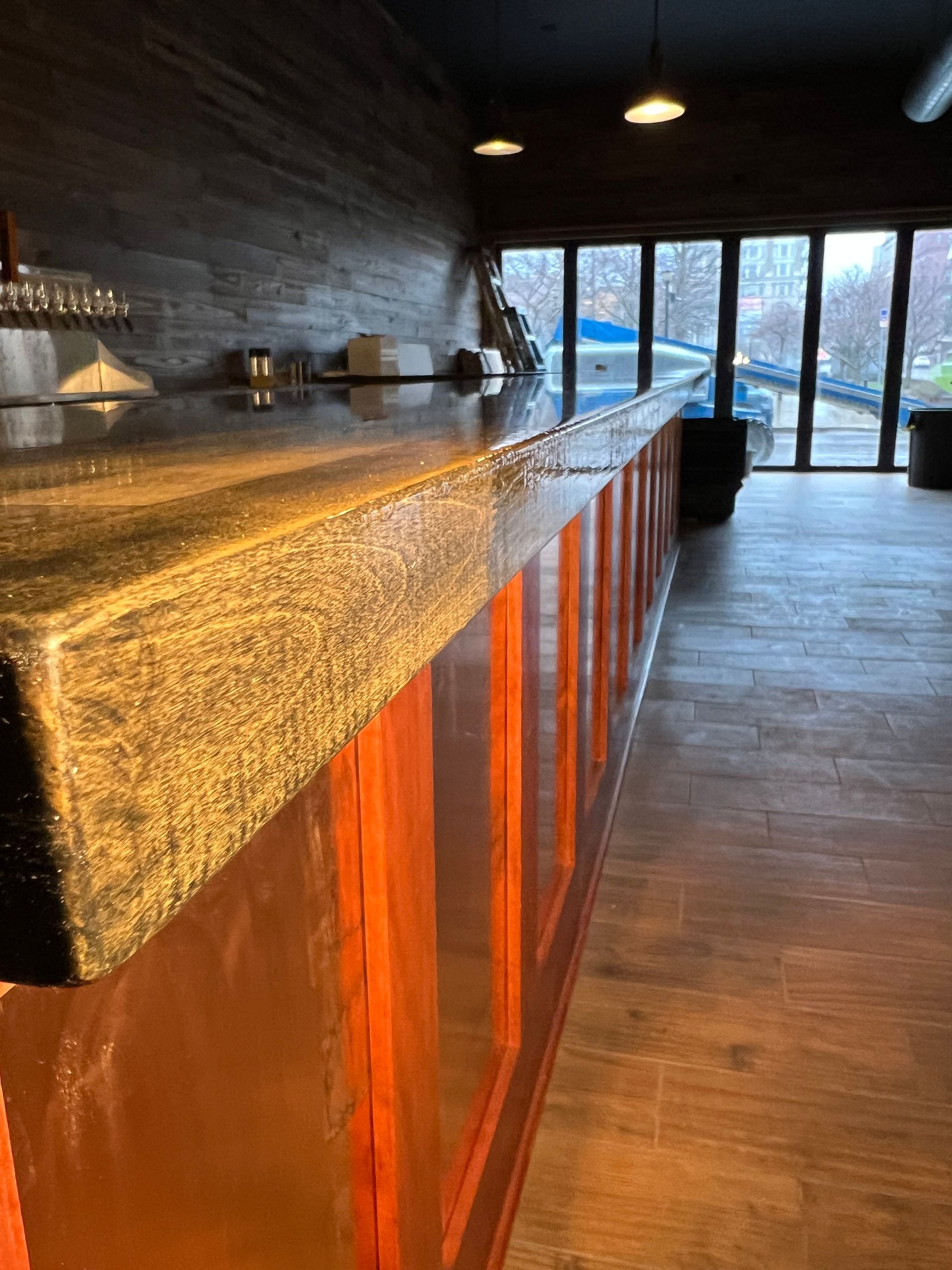 Long wooden bar with dark countertop in a dim room, lit by windows at the far end.
