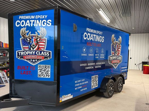 Blue trailer with &ldquo;Premium Epoxy Coatings&rdquo; branding parked indoors, featuring trophy graphics and contact QR code