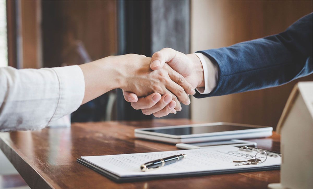Two people shaking hands across a table with paperwork and a pen in an office setting