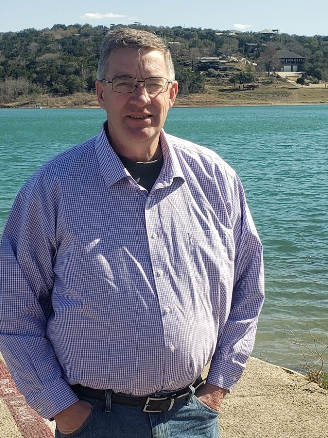 Man standing by a blue waterfront, wearing glasses and a light purple checkered shirt, with hills behind him