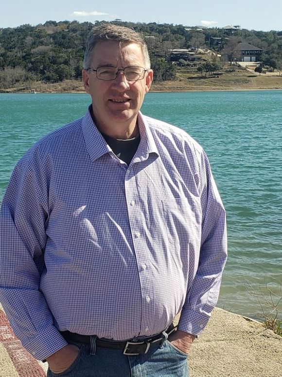 Man standing by a blue waterfront, wearing glasses and a light purple checkered shirt, with hills behind him