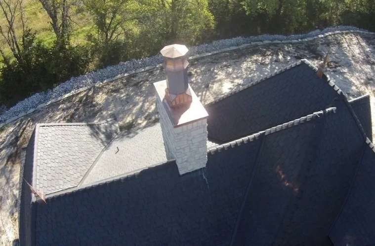 Aerial view of a dark roof with a chimney and slate shingles beside trees.