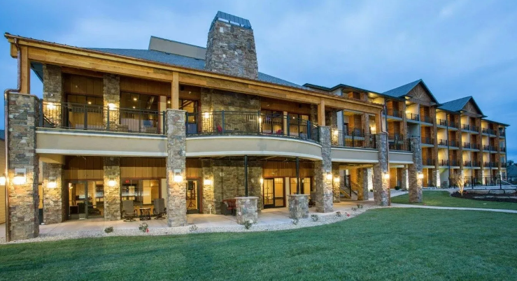 Large rustic lodge with lit balconies and stone chimney at dusk, overlooking a green lawn