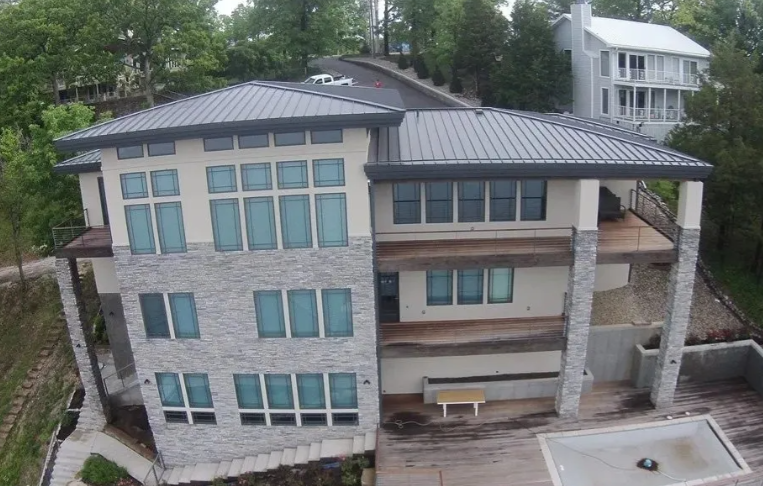 Modern multi-story house with a gray metal roof, large windows, and a covered patio surrounded by trees