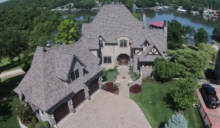 Aerial view of a large stone house with a steep gray roof, trees, driveway, and water behind it.