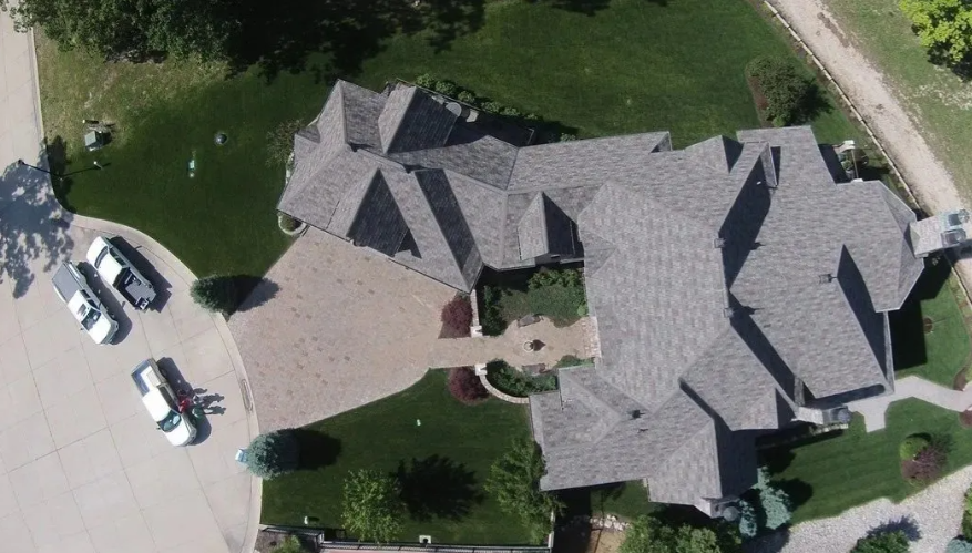 Aerial view of a large gray-roofed house with driveway, parked cars, and landscaped yard